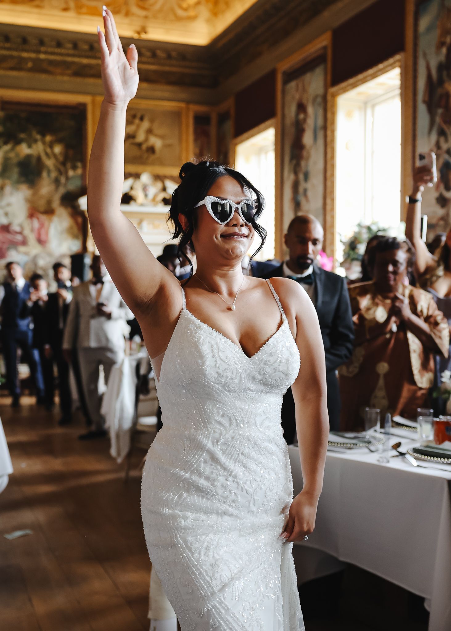 Bride in a white lace dress dances joyfully with raised arms in an ornate ballroom during a wedding reception.