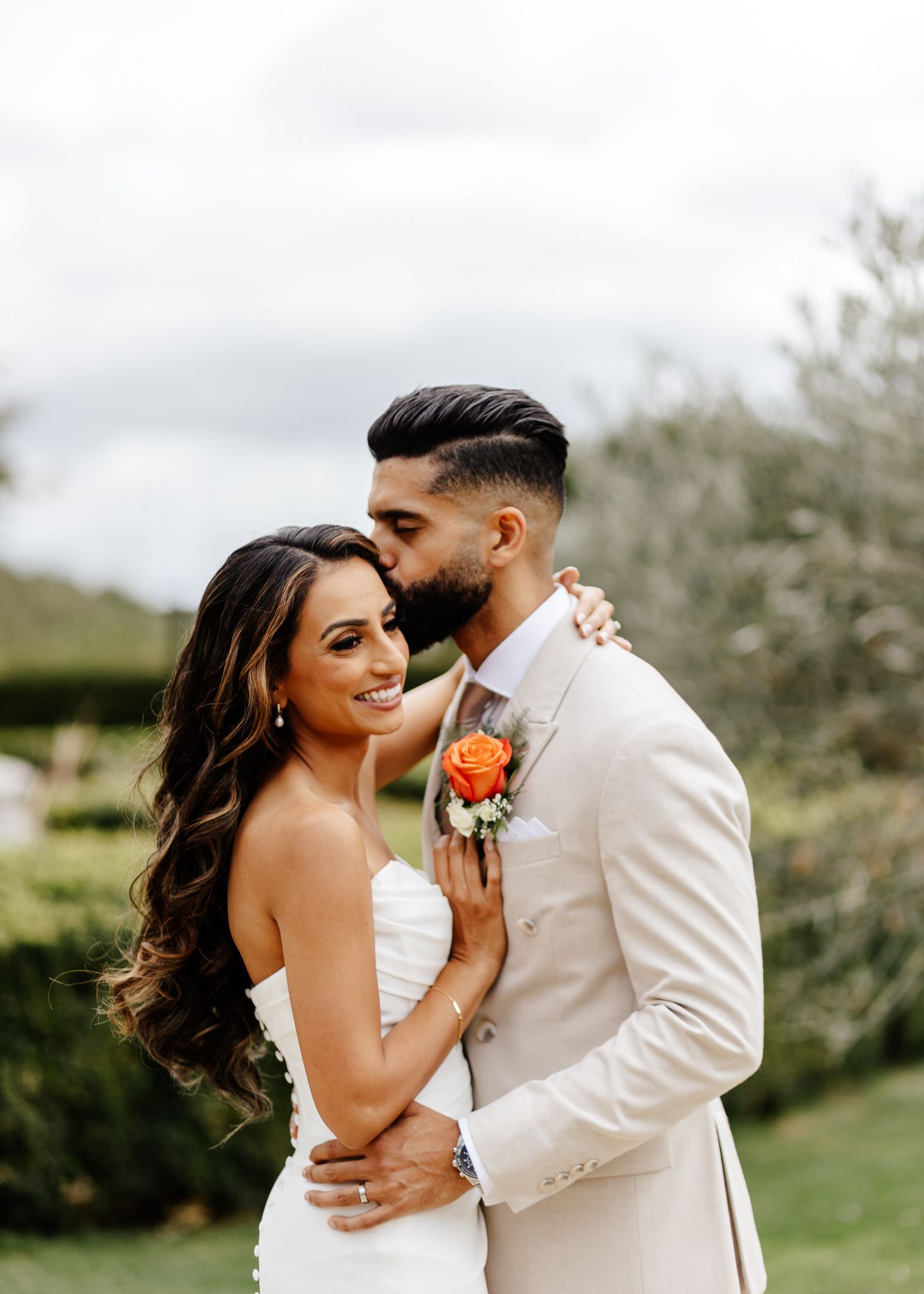 A romantic wedding couple embraces outdoors with the bride in white and groom in cream suit with orange boutonniere.