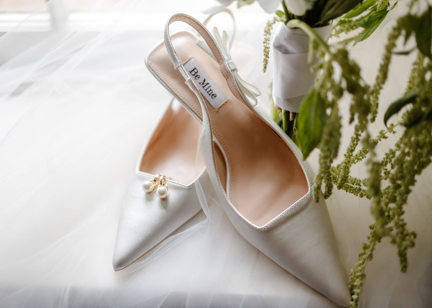 Elegant white satin slingback wedding heels with pearl detail by Badgley Mischka, styled beside green floral accents.
