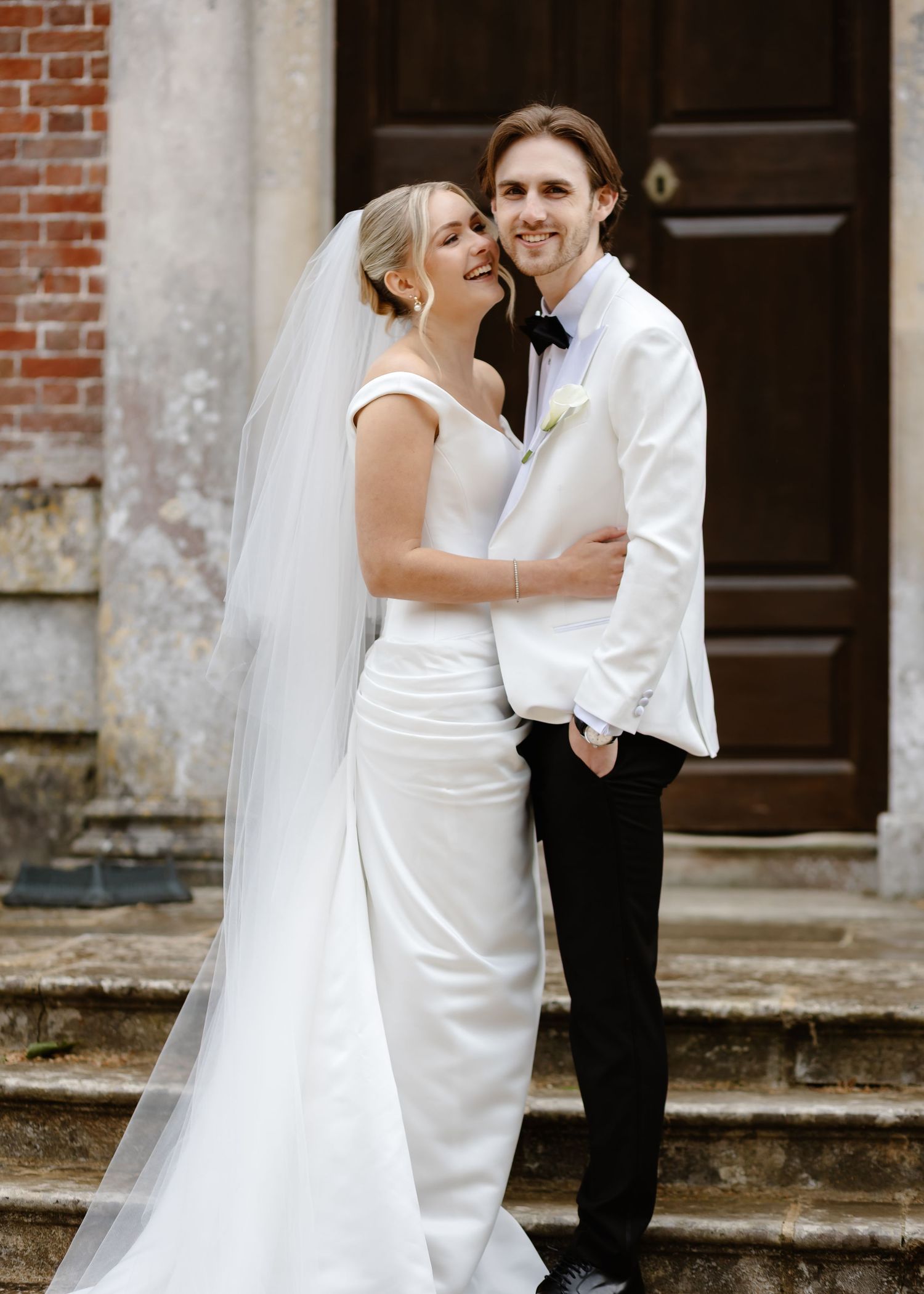 Bride in white gown and veil embraces groom in white jacket and bow tie on stone steps outside a brick building.