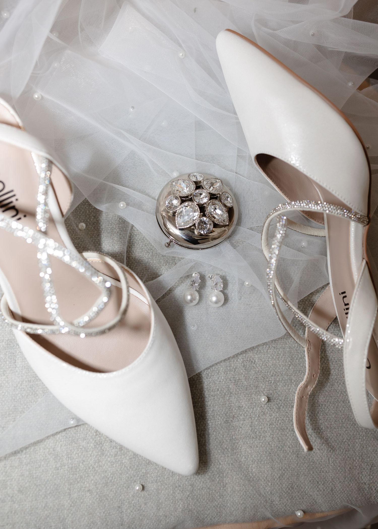 Elegant white bridal heels with pearl straps beside crystal jewelry and stud earrings on a soft gray surface.