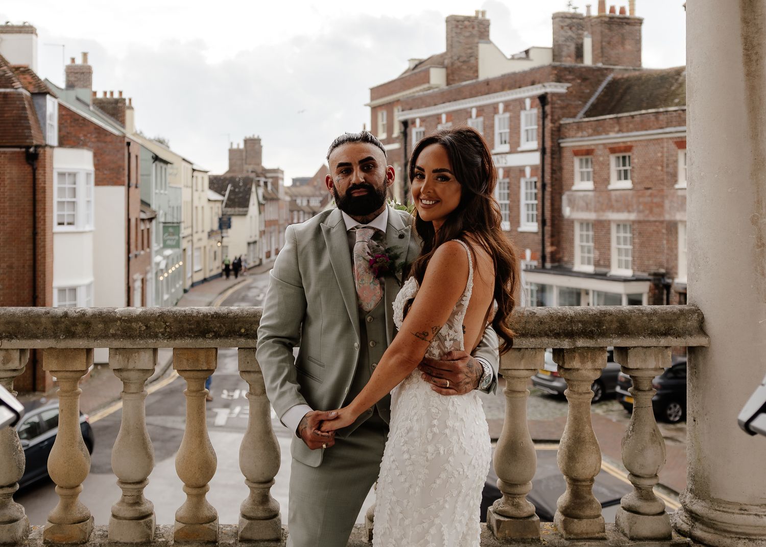 Wedding couple embraces on a balcony overlooking historic brick buildings on their special day.