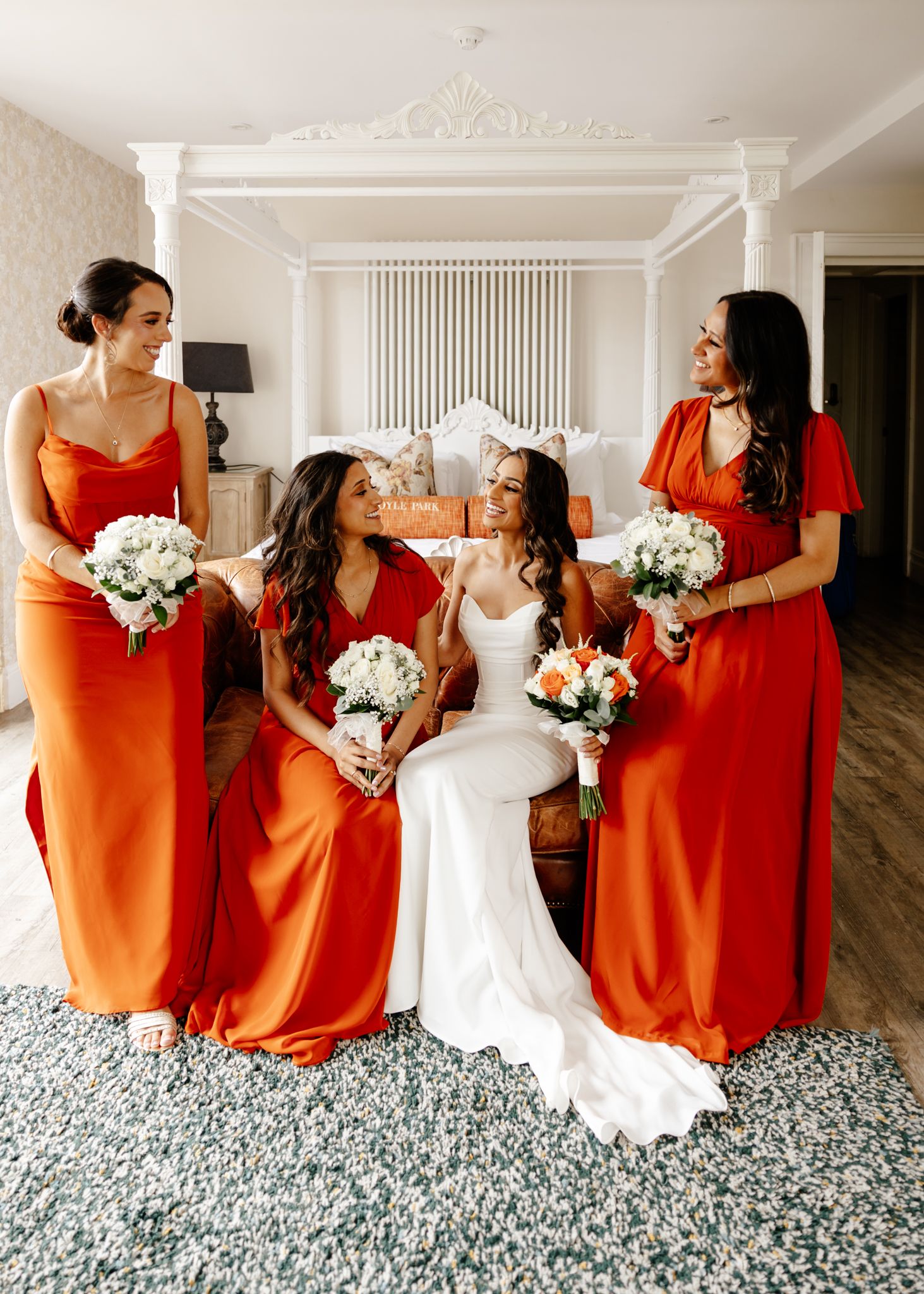 A wedding party in bright orange bridesmaid dresses poses together holding white bouquets on a patterned carpet.