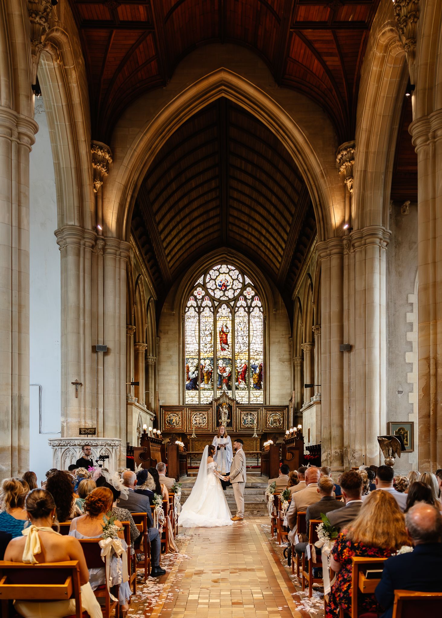 Wide panoramic view of a traditional church wedding ceremony with guests seated in wooden pews under vaulted Gothic arches.