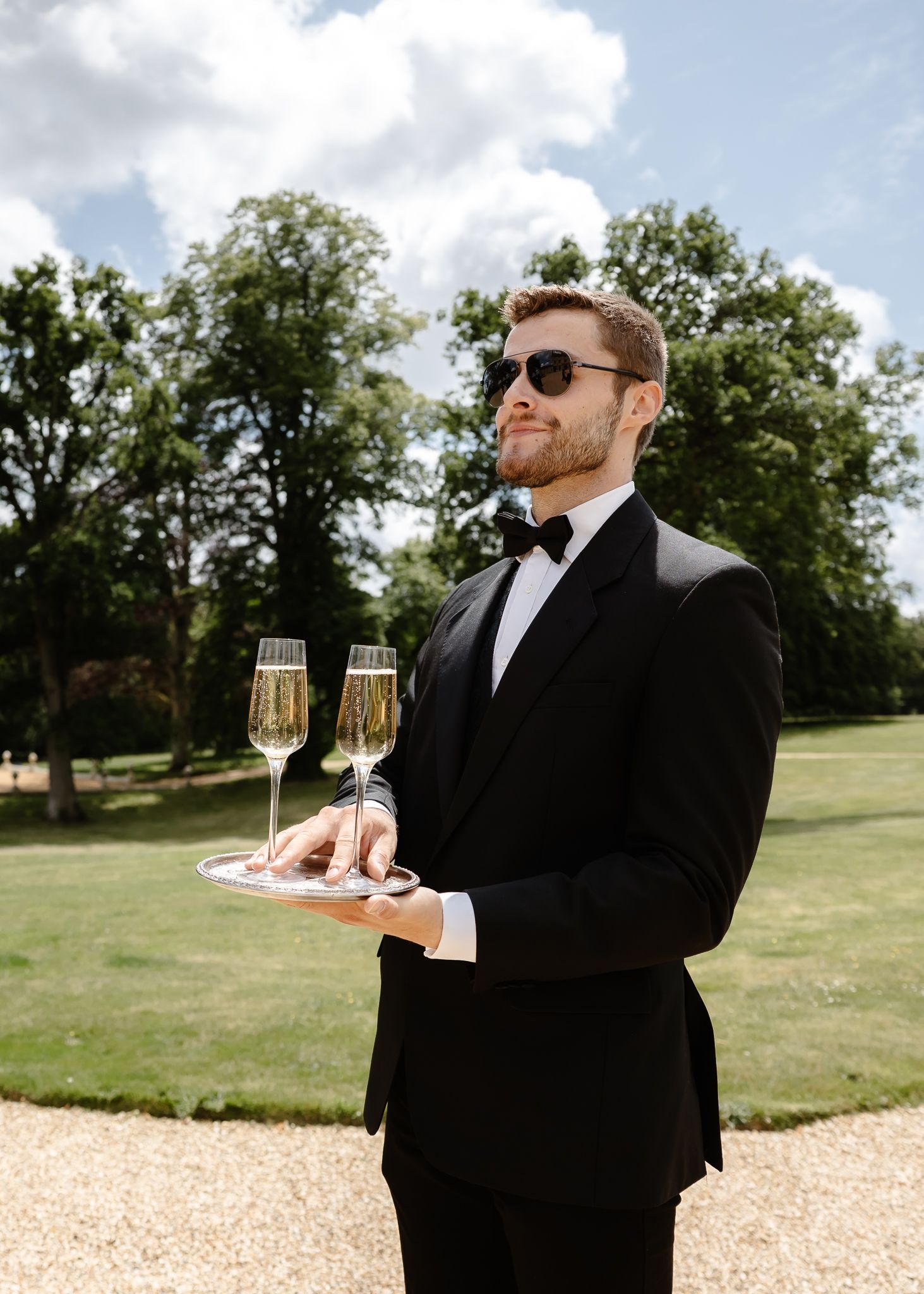 A groomsman in a black tuxedo and sunglasses carries champagne flutes on the lawn of an elegant estate.
