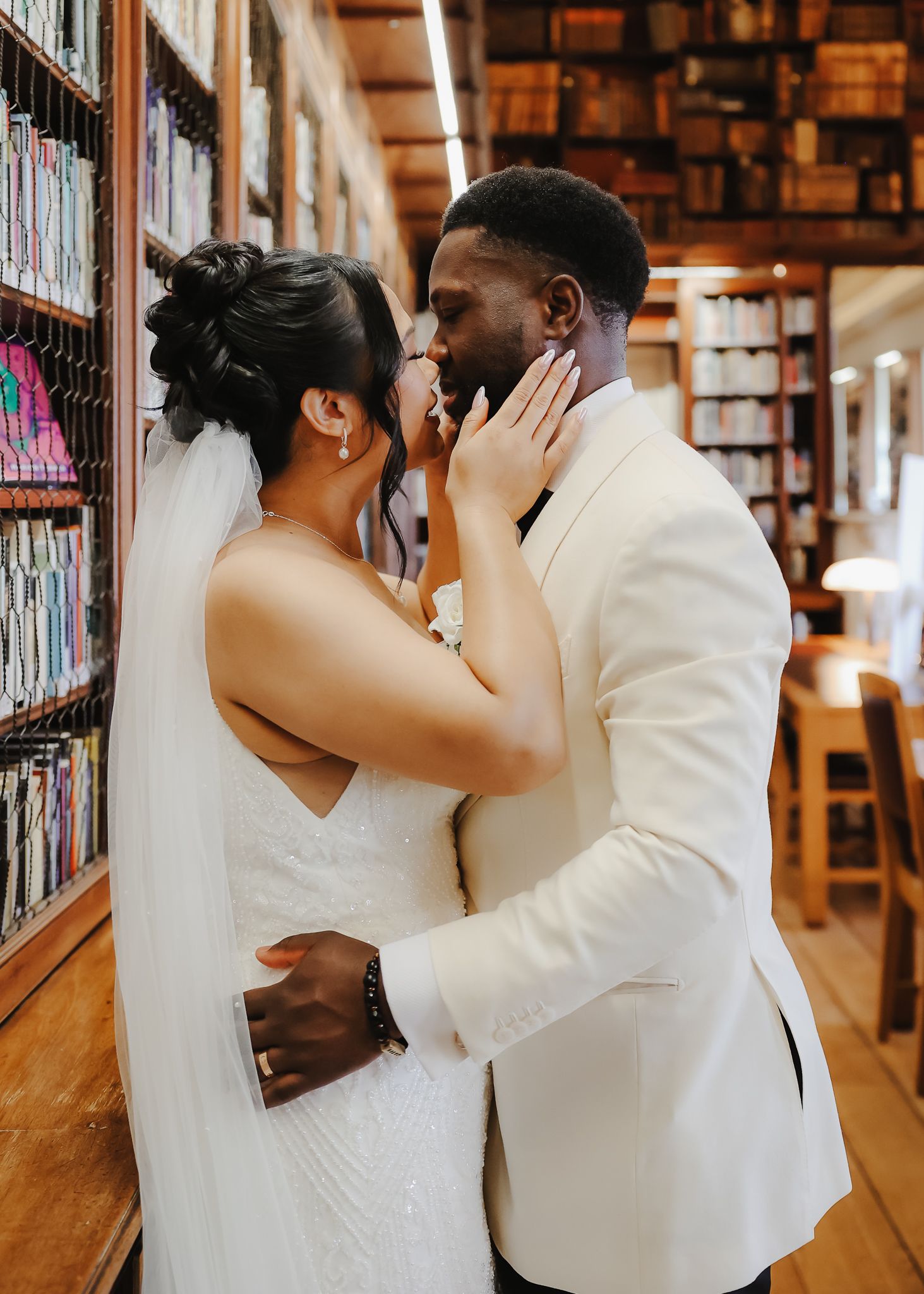 A couple in wedding attire share an intimate moment between bookshelves in a library setting.