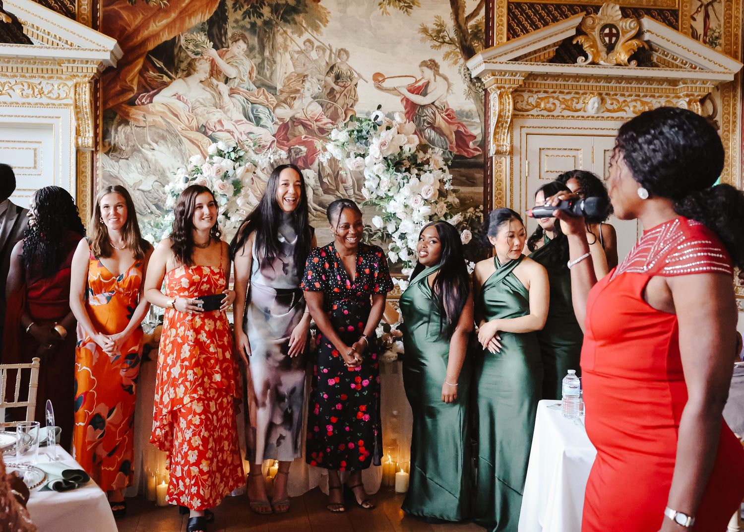 Wedding guests in colorful dresses stand together in front of an ornate floral display at an elegant venue.