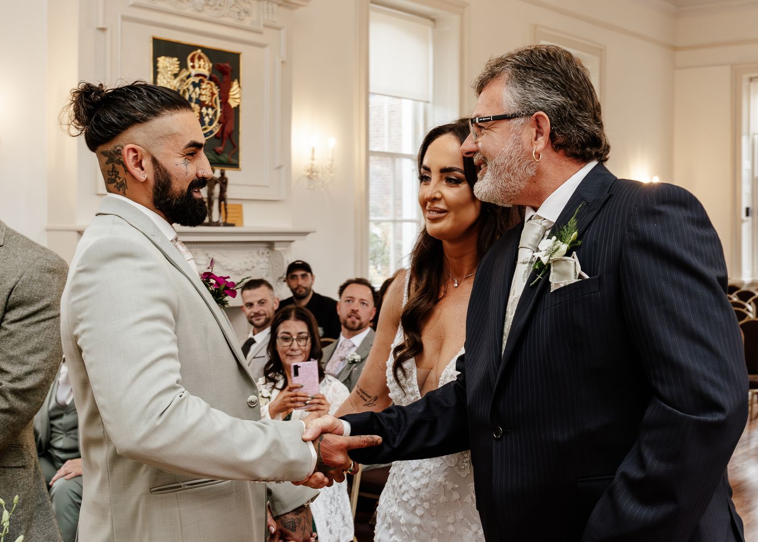 Wedding ceremony guests gather in an elegant white room with ornate crown molding and wall sconces.