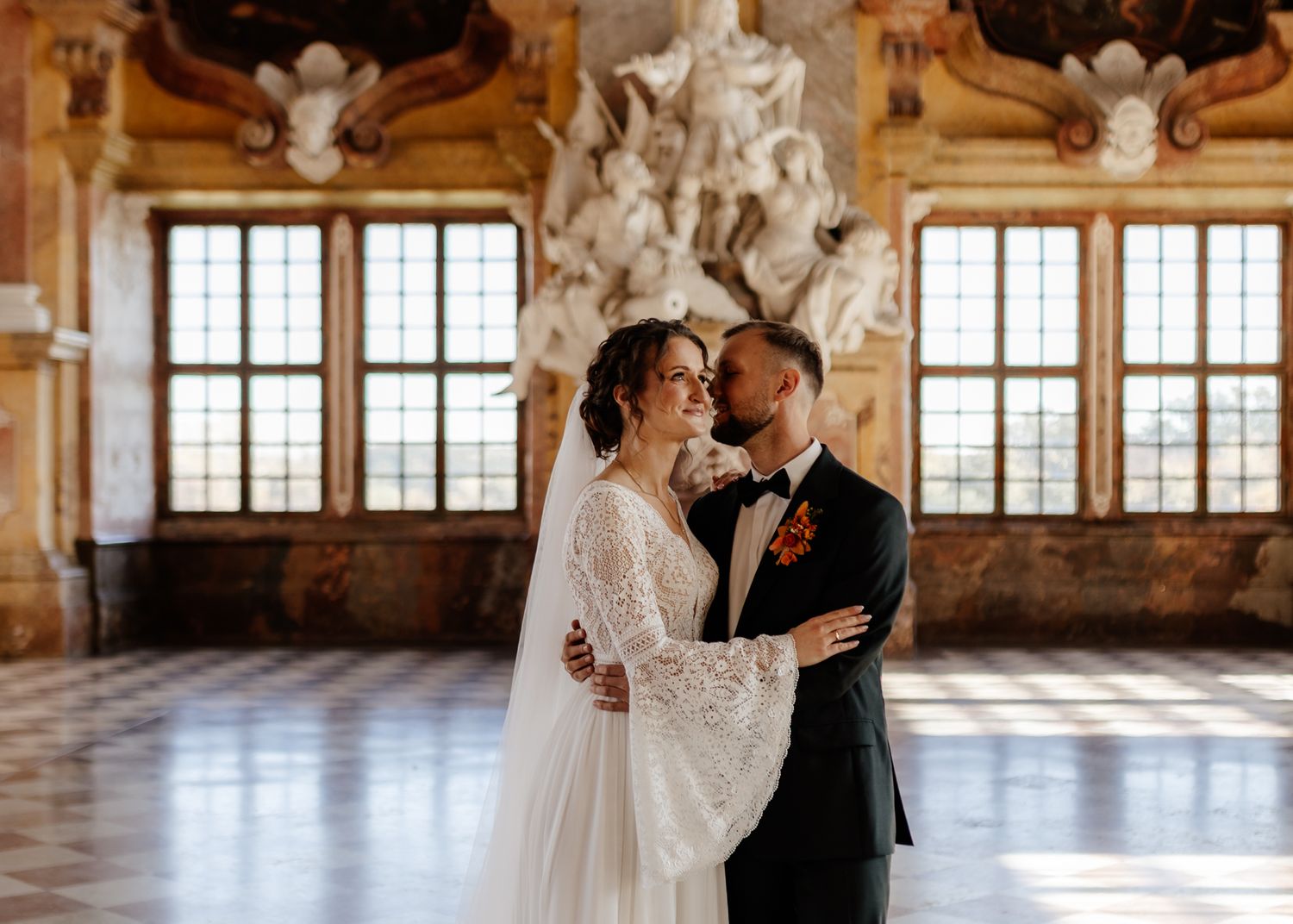 A couple shares intimate moments in an elegant ballroom with ornate architecture and large windows.