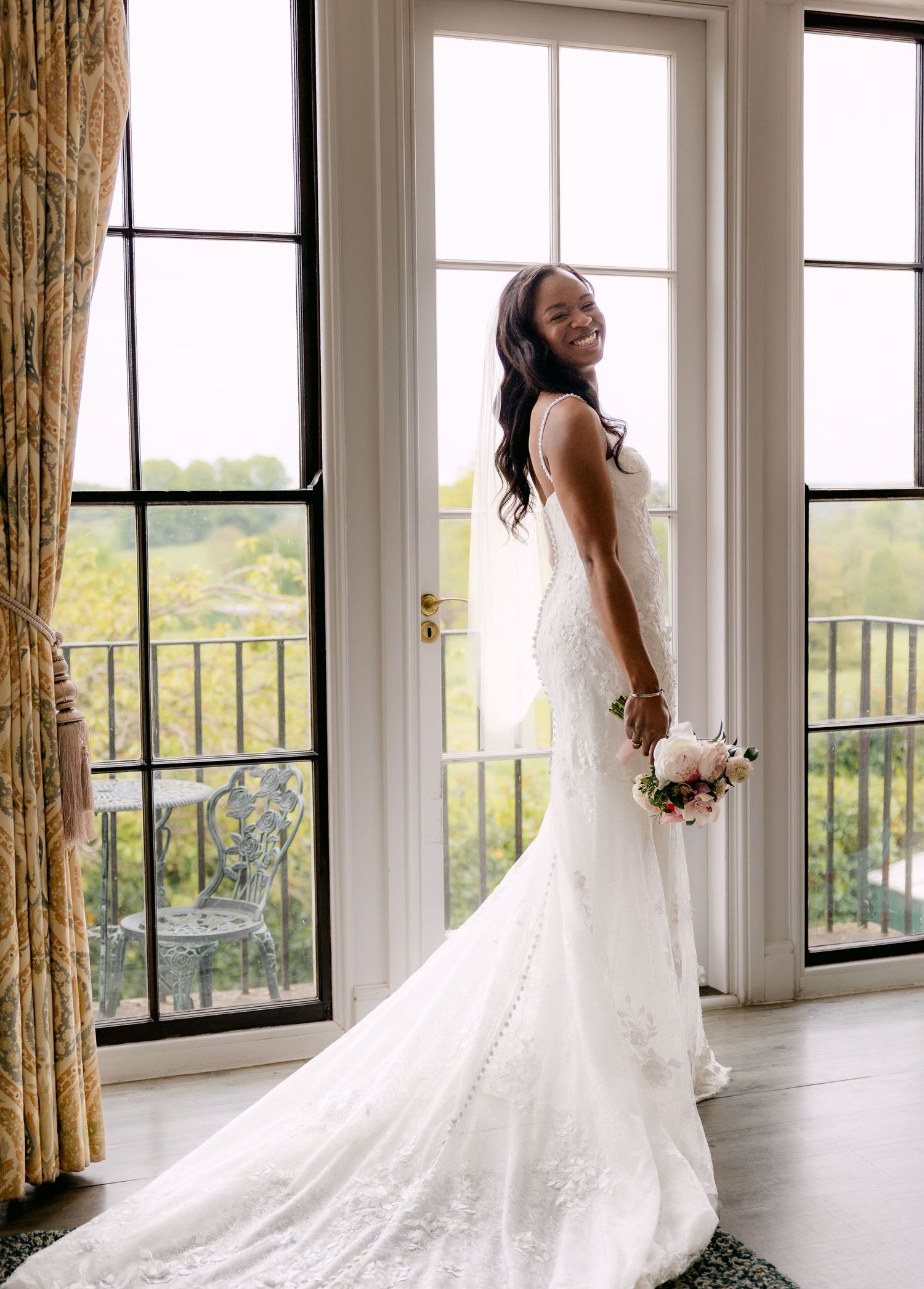 Smiling bride in white lace mermaid gown with train holds bouquet by French doors with garden view.