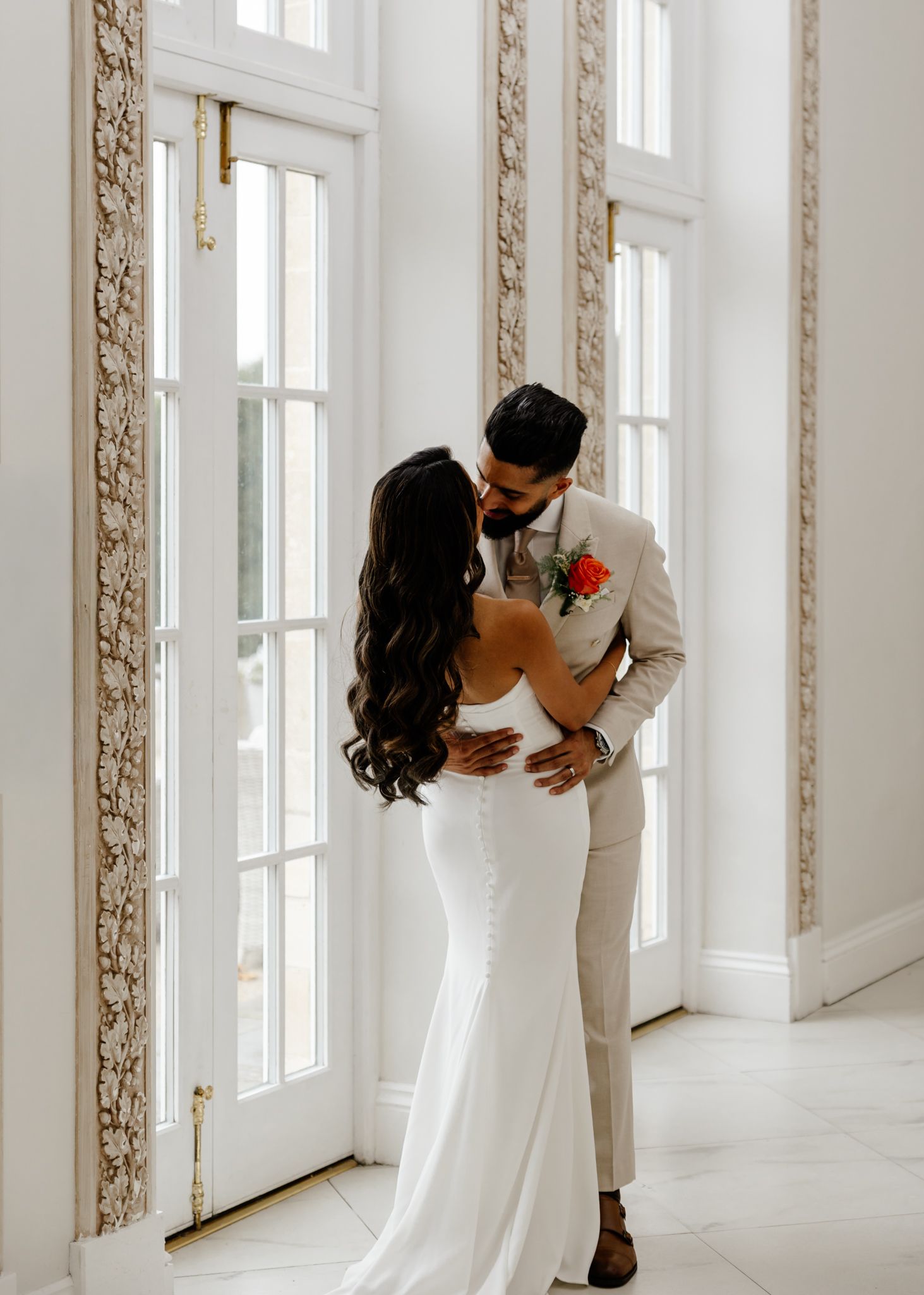 Elegant couple in white attire share an intimate moment by tall ornate windows in a bright wedding venue.