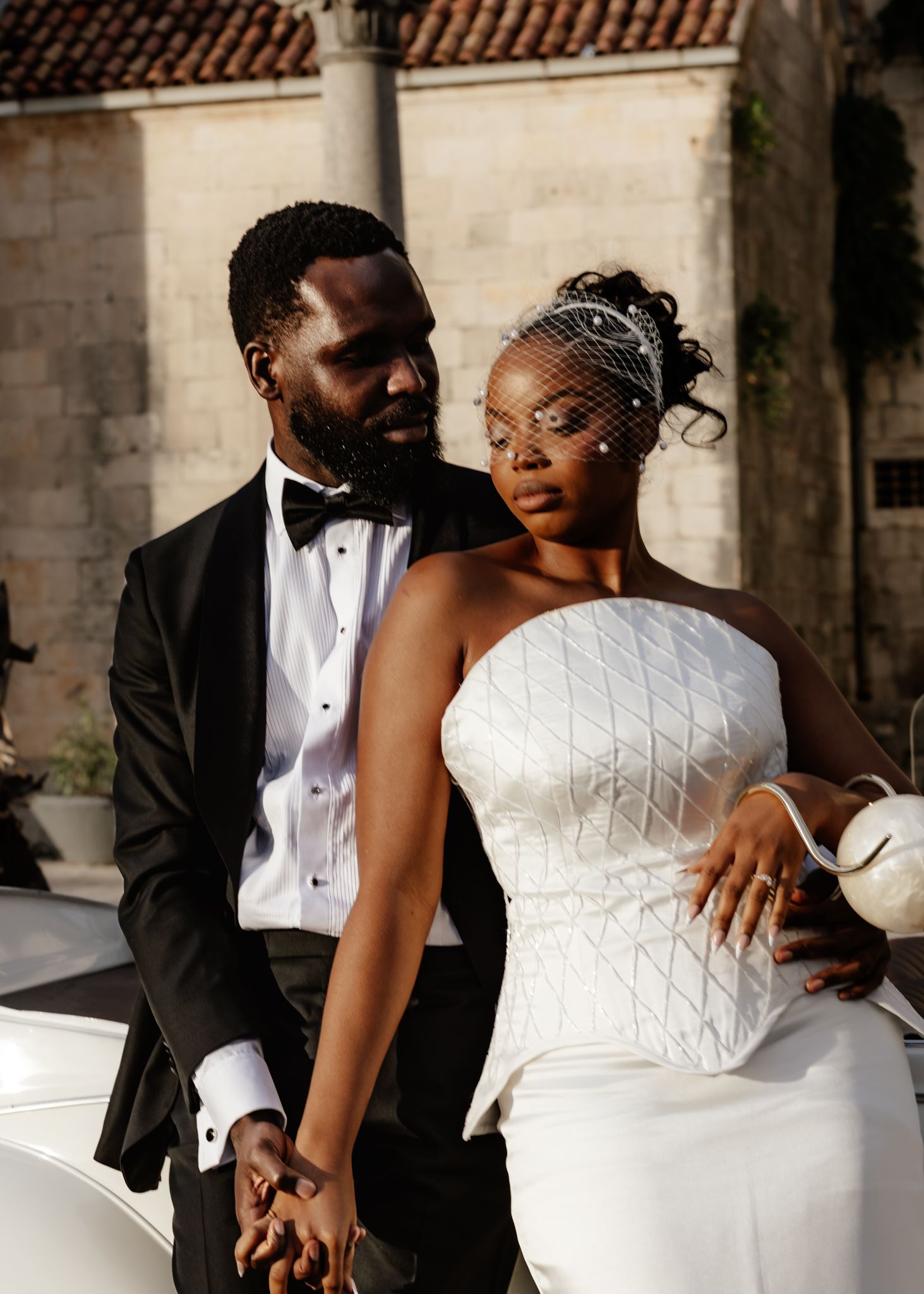 Elegant couple in formal wedding attire pose together outdoors against stone architecture with tiled roof.