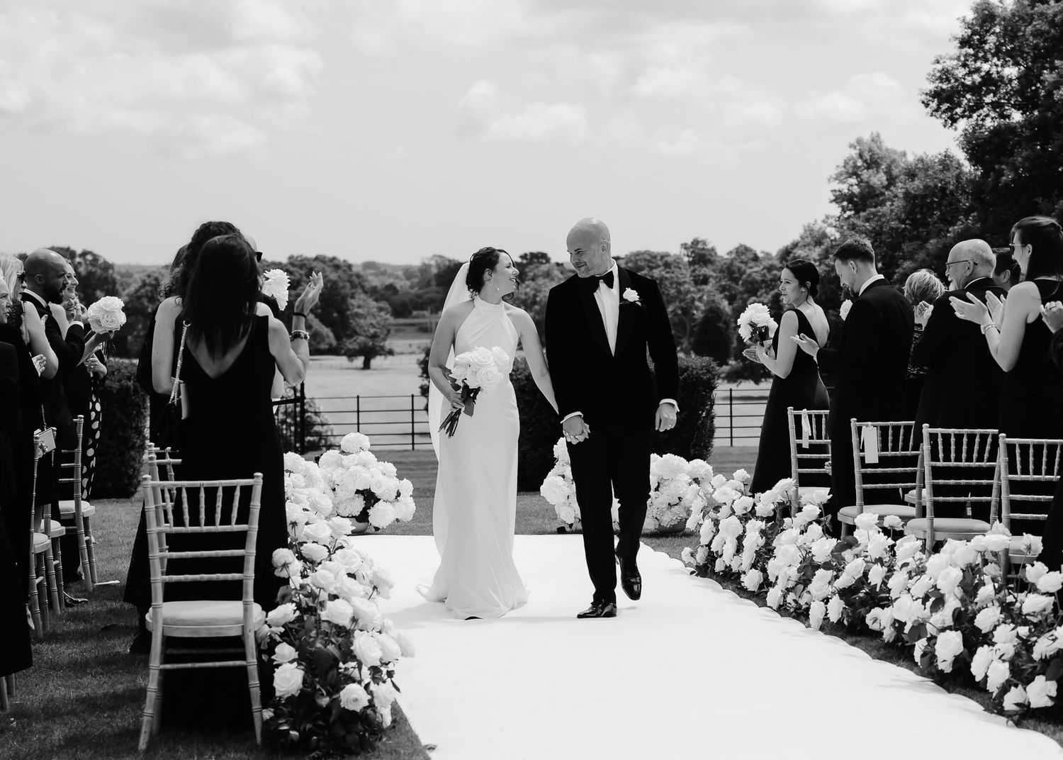 Outdoor wedding recessional on white aisle runner lined with lush white floral arrangements.