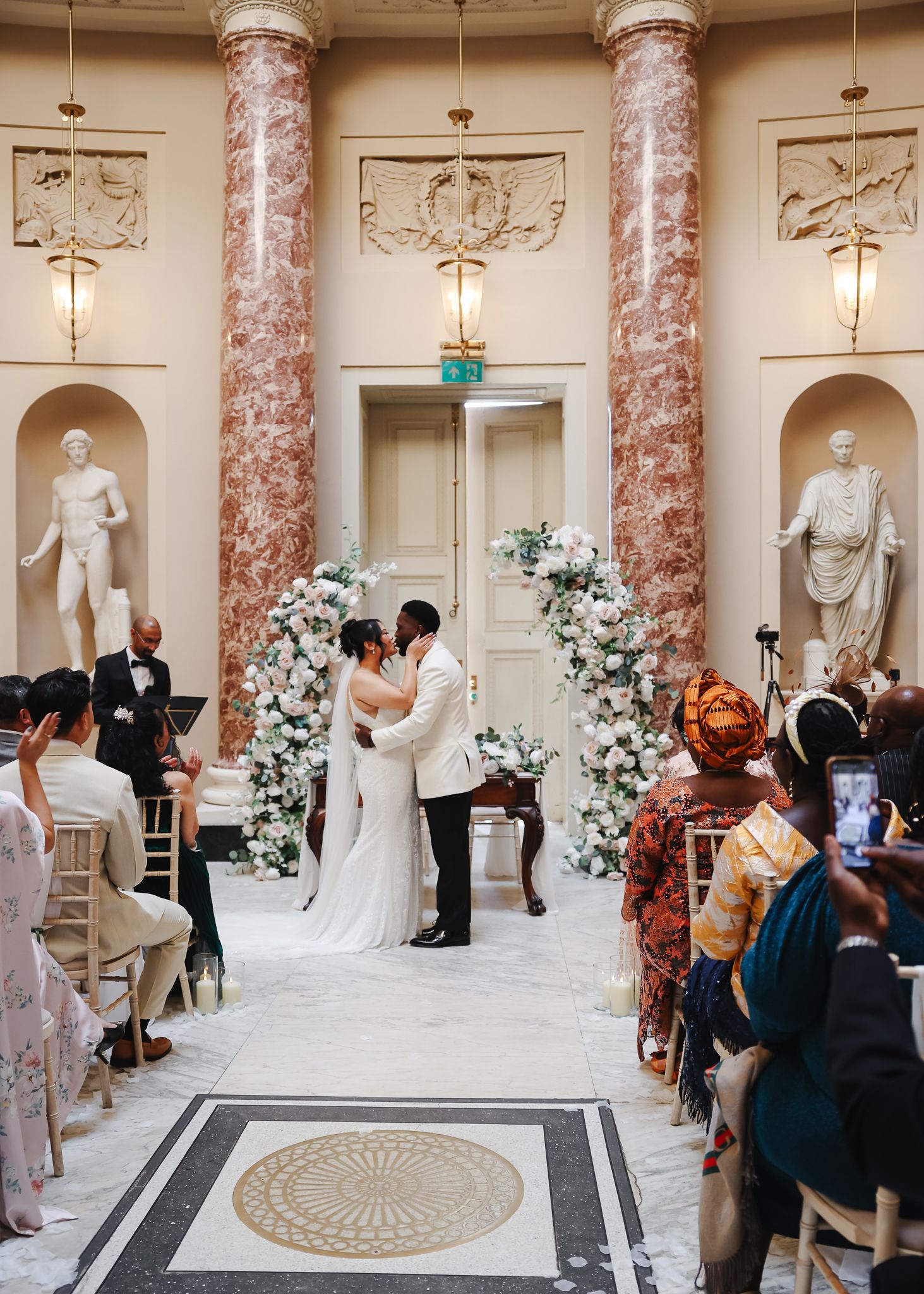 Elegant wedding ceremony in a grand marble hall with pink columns, white floral arrangements, and classical statues.