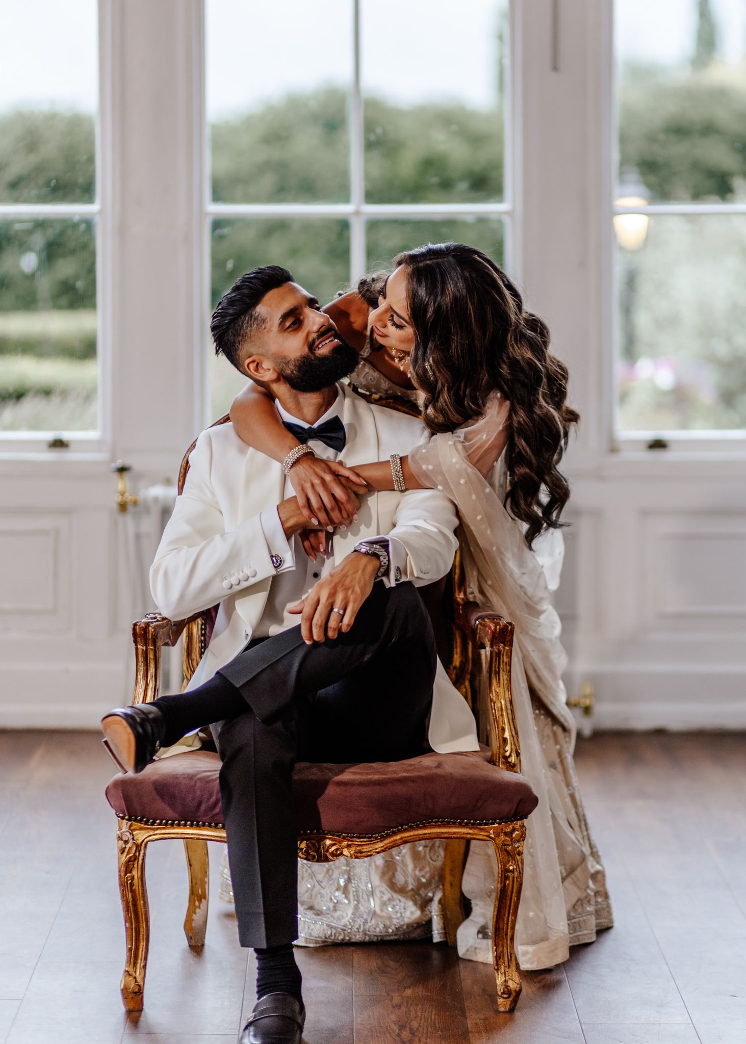 A couple shares intimate moments on an elegant vintage chair in a bright sunlit room with large windows.