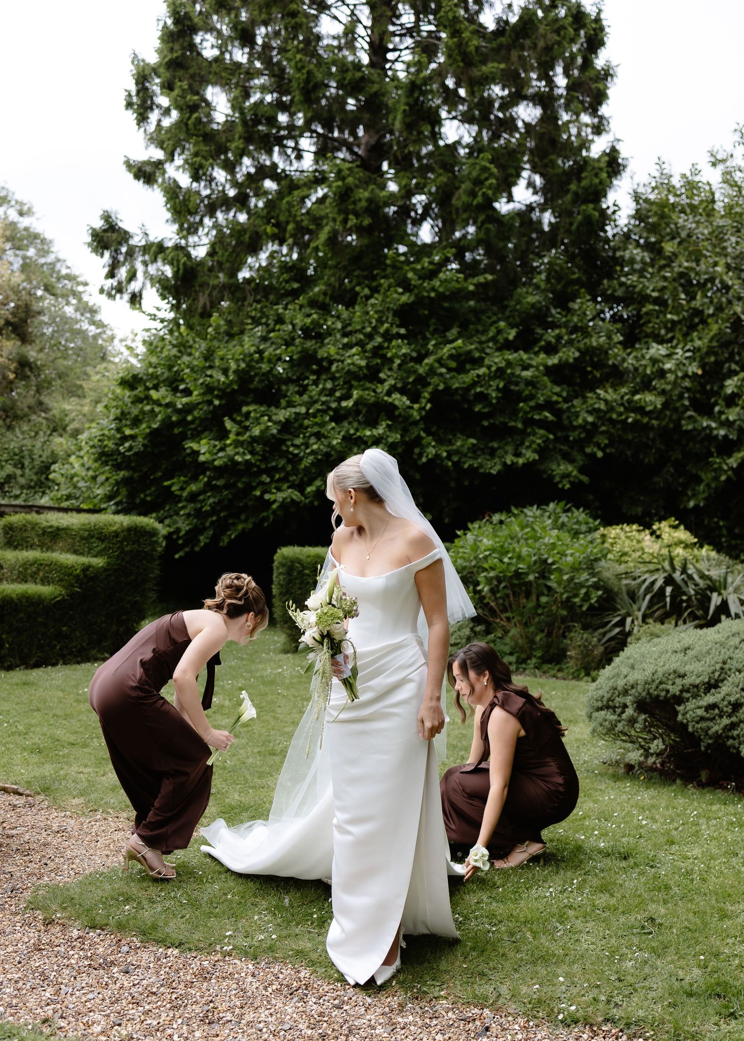 Bride in white gown holds bouquet while two bridesmaids in brown dresses adjust her dress train in a lush garden.
