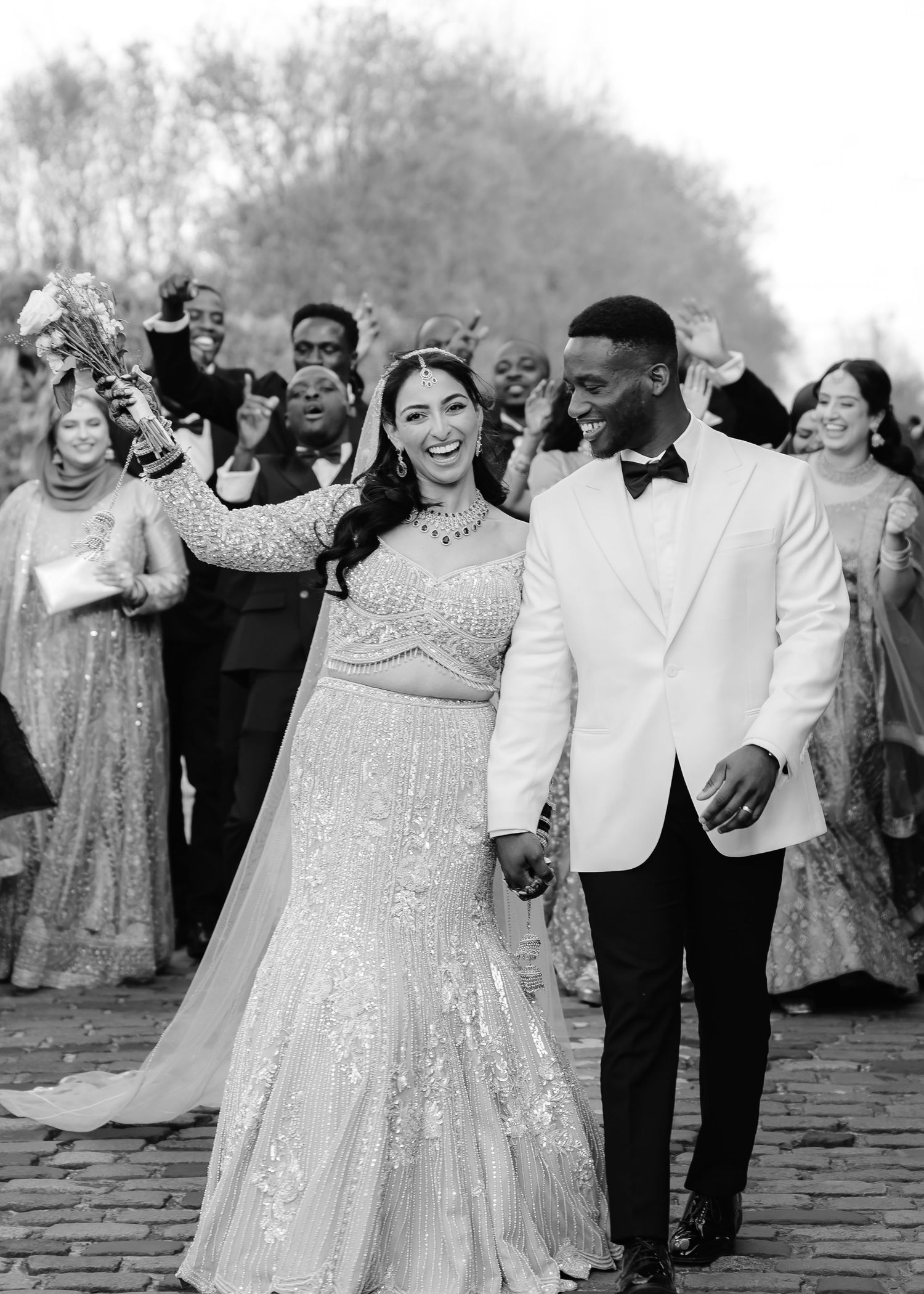 Black and white photo of a joyful couple in wedding attire holding hands, surrounded by celebrating guests outdoors.