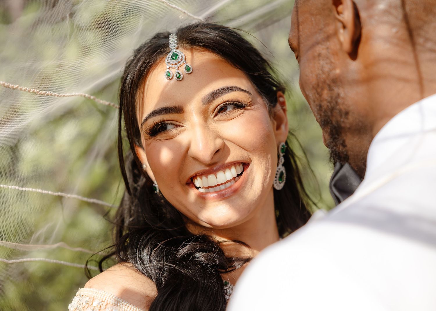 Smiling South Asian bride in traditional jewelry and maang tikka gazes lovingly at groom during outdoor wedding ceremony.