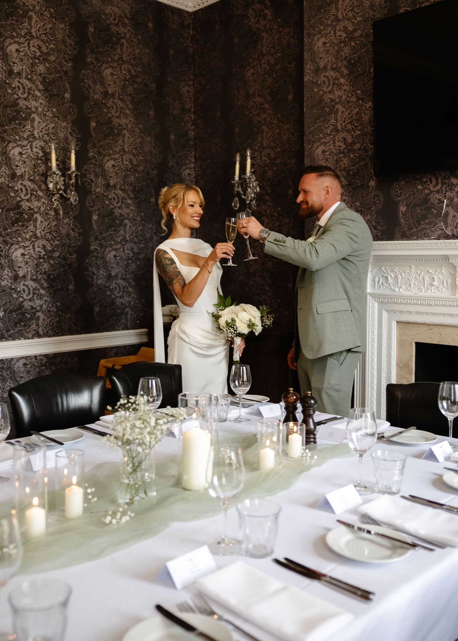 Bride and groom toasting champagne at elegantly set wedding reception table in a sophisticated dark-wallpapered dining room.