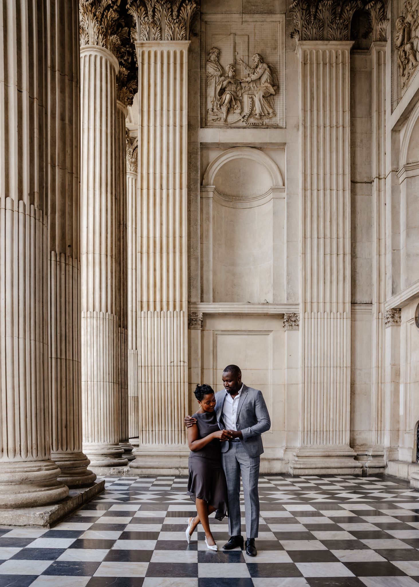A couple shares an intimate moment in a grand hallway with classical columns and checkerboard marble floors.