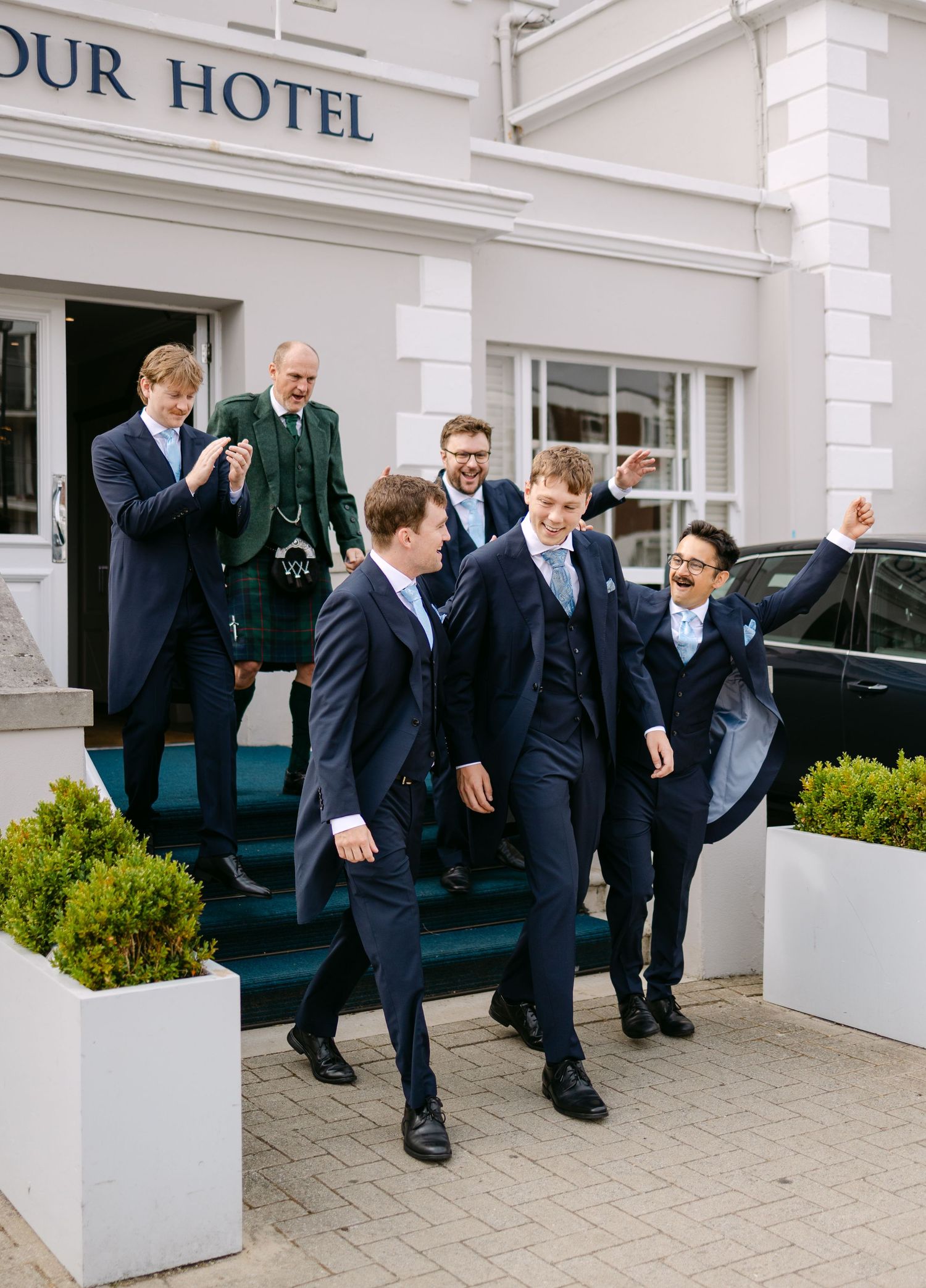 Groomsmen in navy suits laugh outside a hotel entrance, with one man in a green Scottish kilt on the steps.
