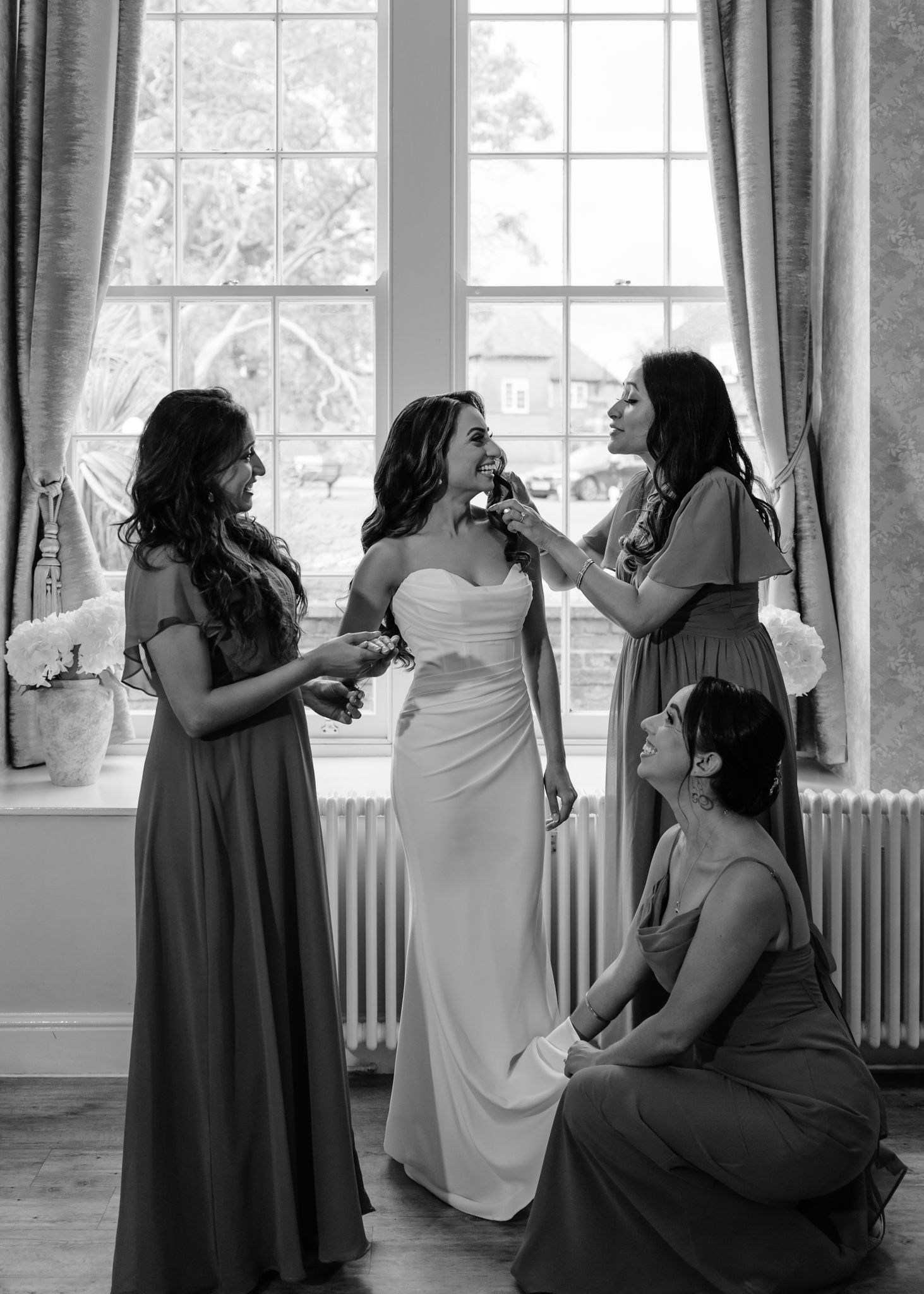 Wedding party getting ready in a bright room with large windows and gray curtains.