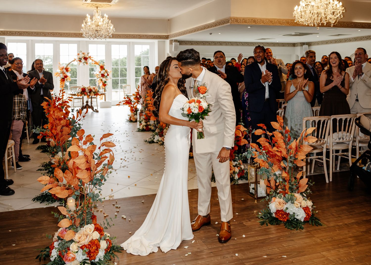 Wedding reception venue decorated with orange florals, crystal chandeliers, and white chairs.