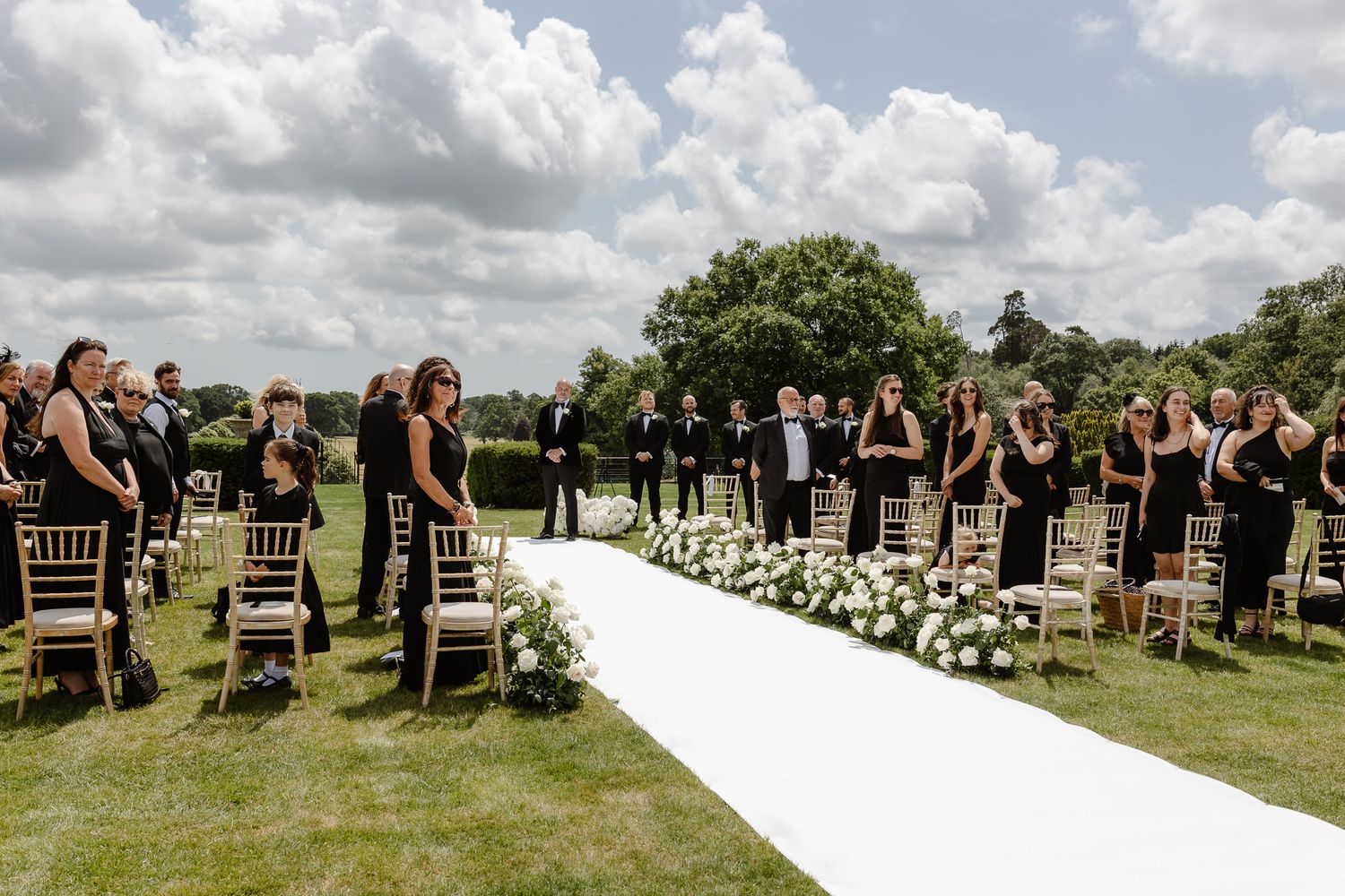 Outdoor wedding ceremony setup with rows of wooden chairs on a grassy lawn under cloudy skies.