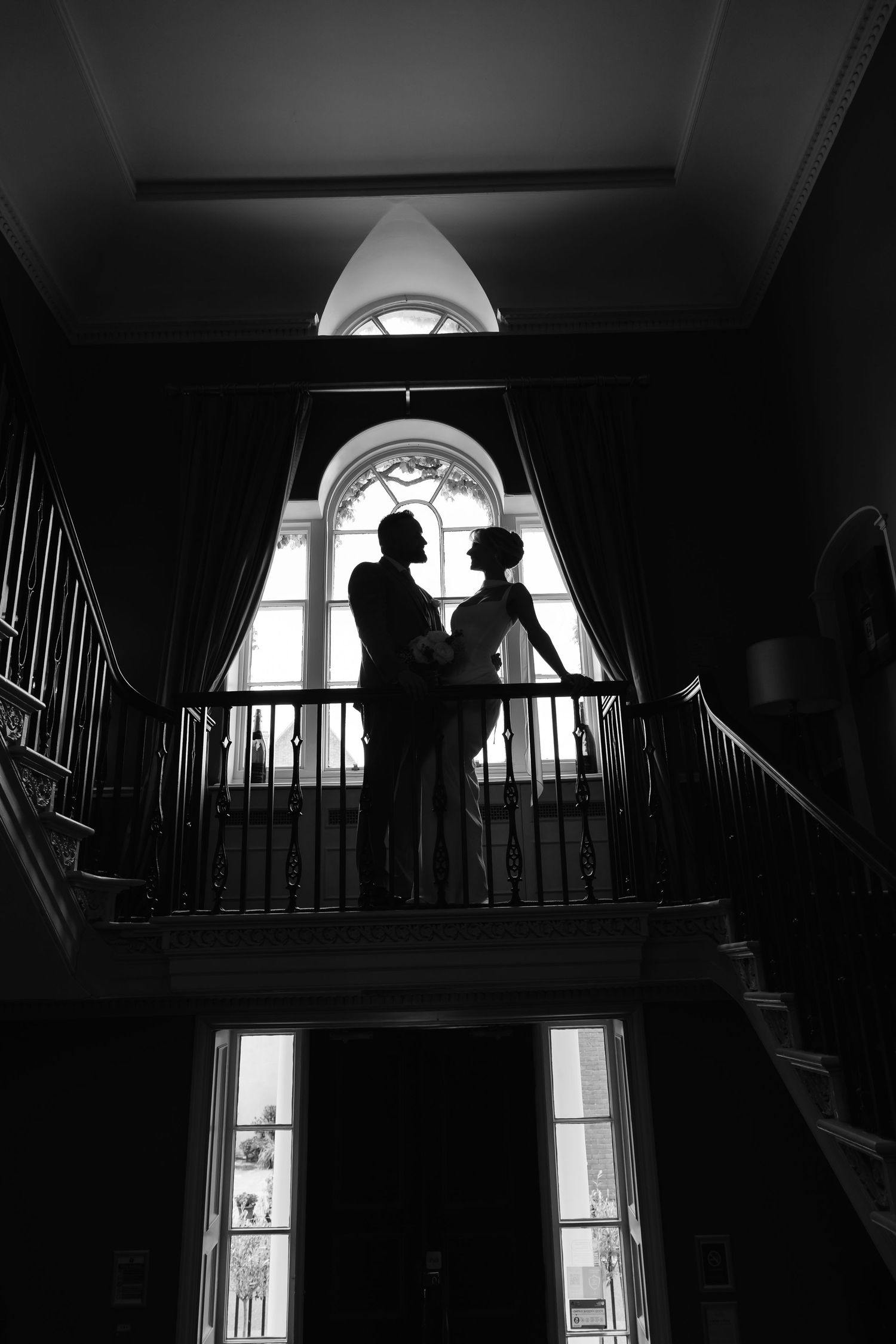 Silhouette of couple standing on balcony in front of arched window in elegant interior with dramatic black and white lighting.