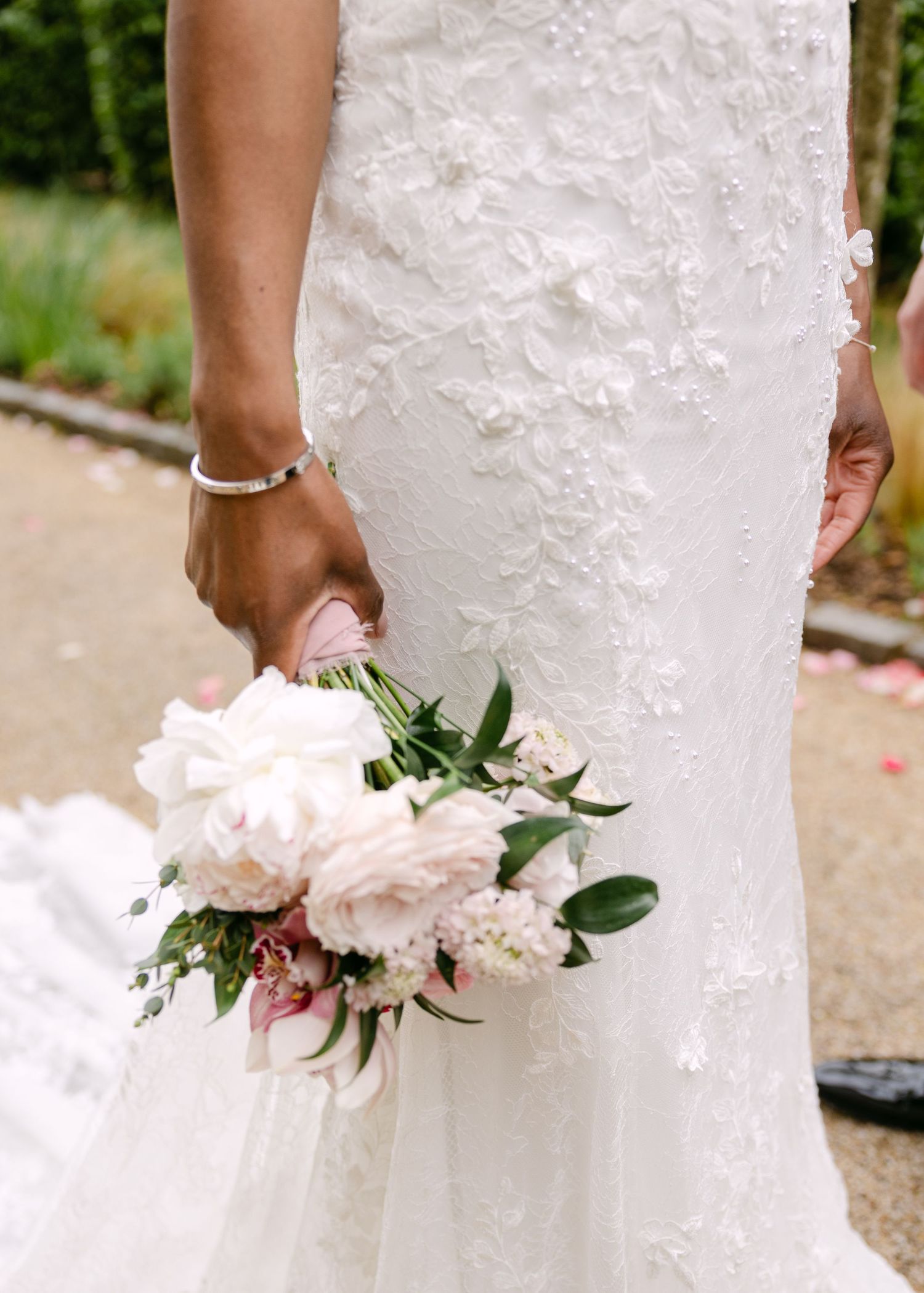 Bride in white lace wedding gown holding a bouquet of blush pink peonies and greenery at an outdoor wedding.