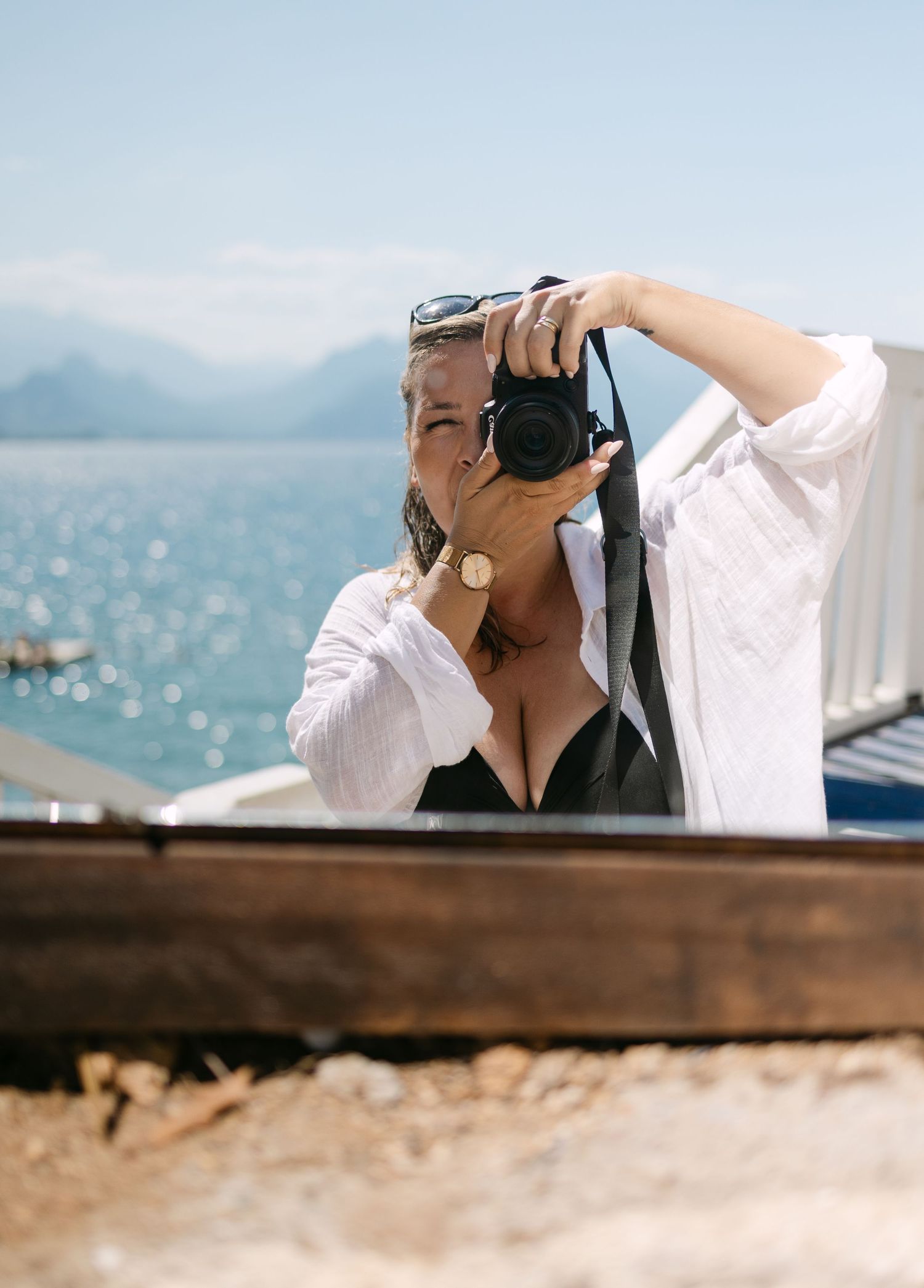 Woman in white shirt photographs with DSLR camera outdoors near sparkling blue lake with mountain backdrop on sunny day.