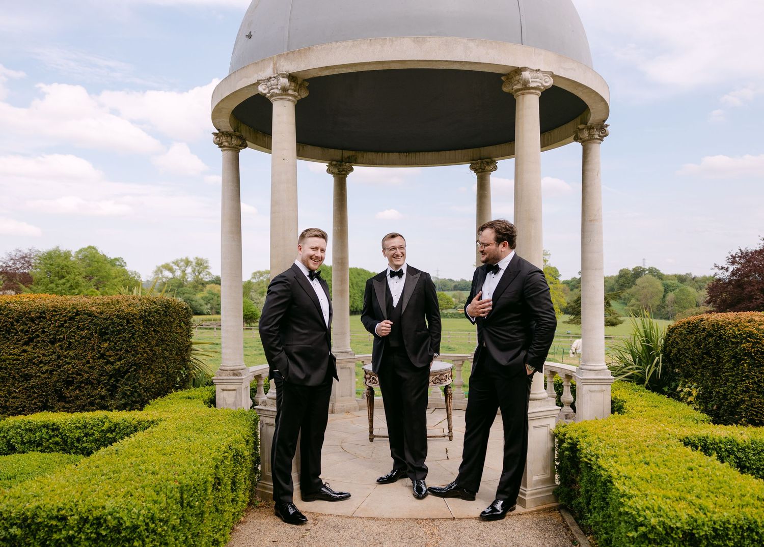 Three groomsmen in black tuxedos stand beneath an elegant garden gazebo surrounded by manicured hedges.