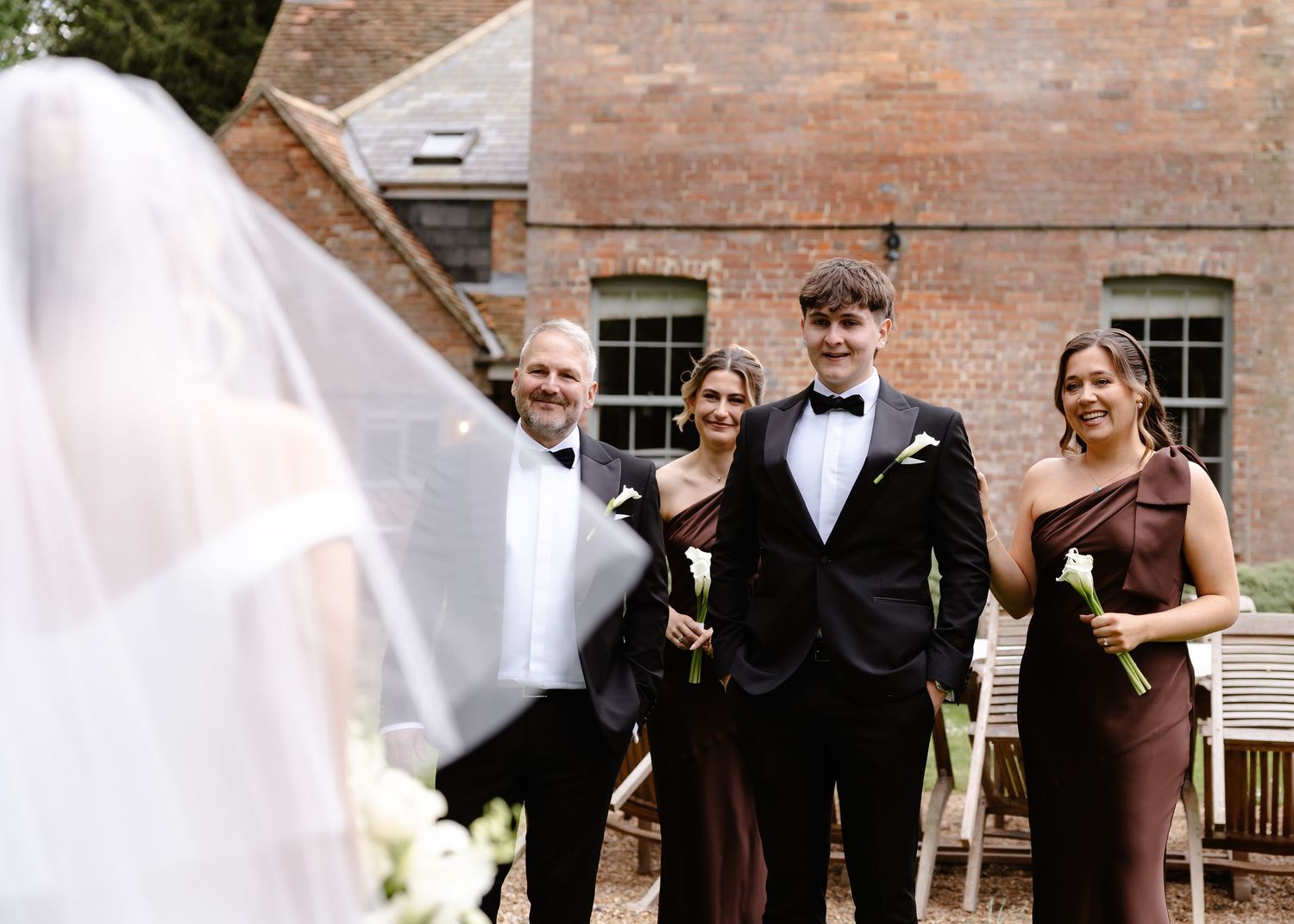 Groom in black tuxedo smiles at veiled bride, flanked by groomsmen and bridesmaids in burgundy at outdoor wedding.