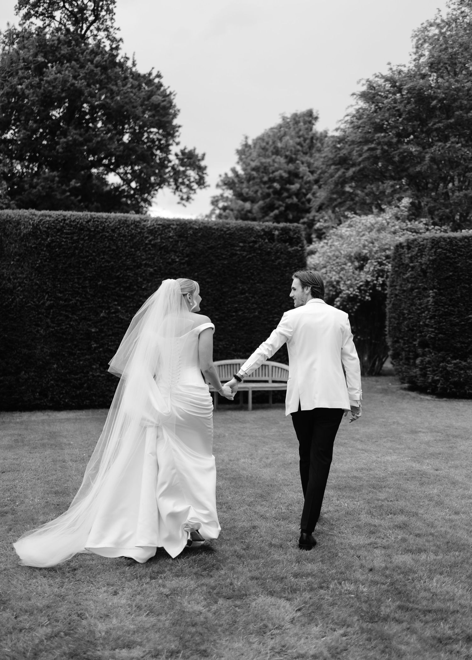 Bride and groom walk hand in hand across a manicured lawn, captured in elegant black and white photography.