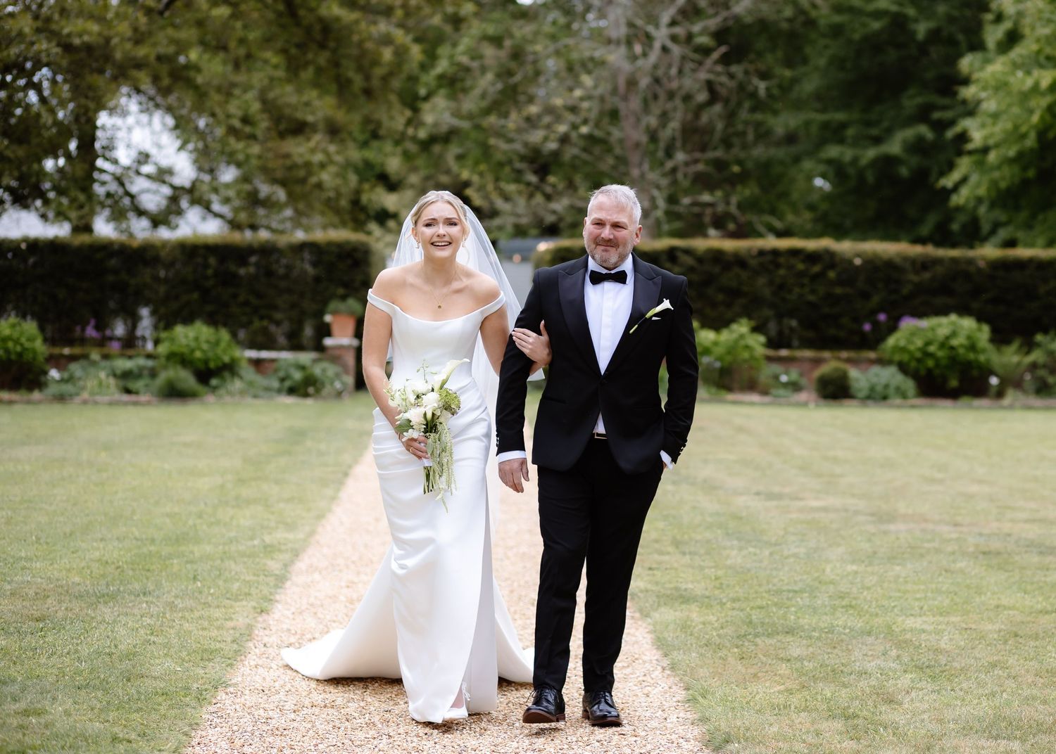 Bride in white off-shoulder gown holding bouquet walks with suited man on garden path at elegant outdoor wedding.