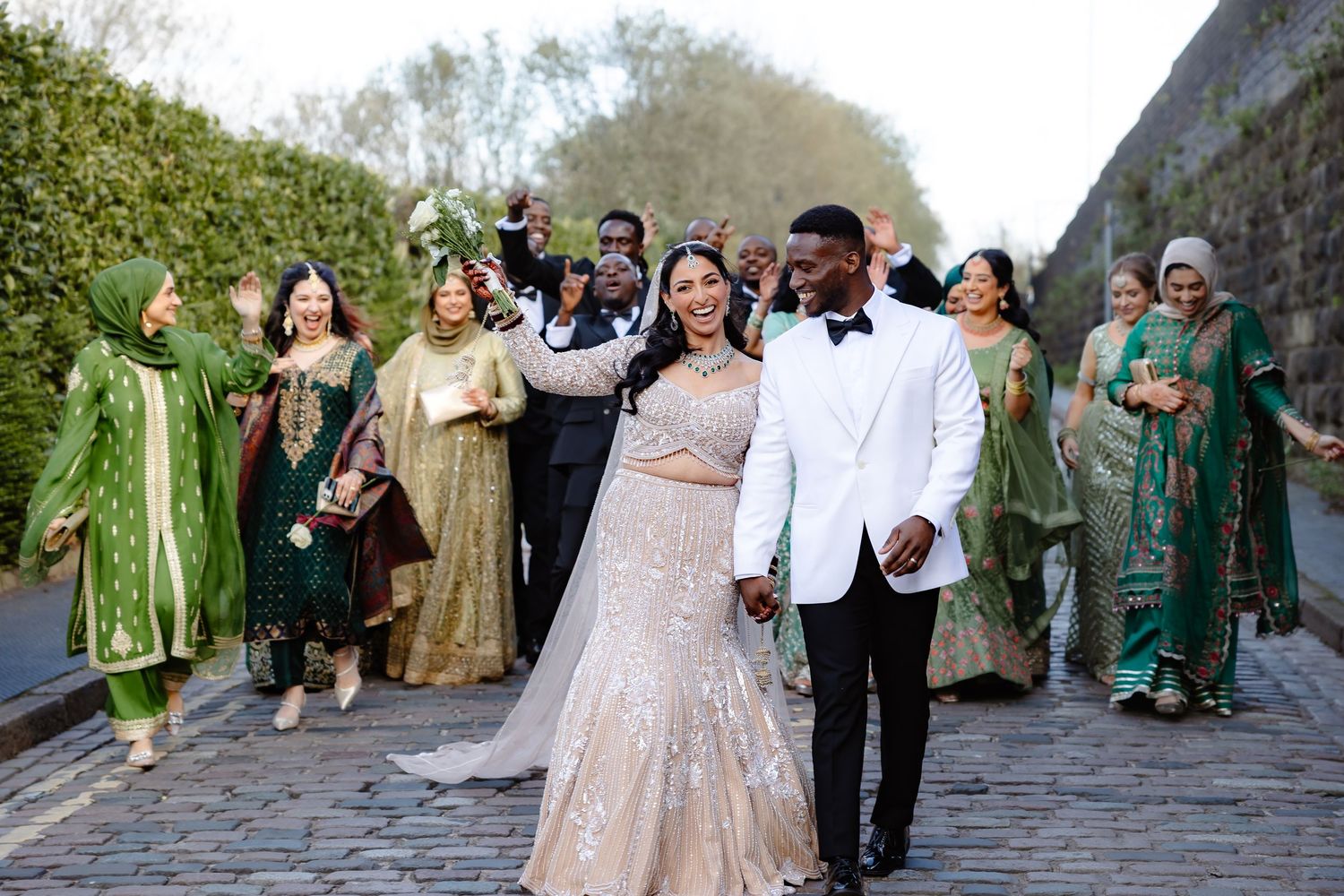 A joyful wedding party celebrates outdoors, with the bride in a gold gown and groom in a white tuxedo leading the group.
