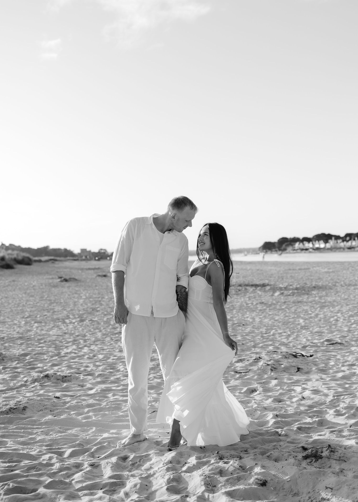 Black and white photo of a couple in white attire sharing a romantic moment on a sandy beach.