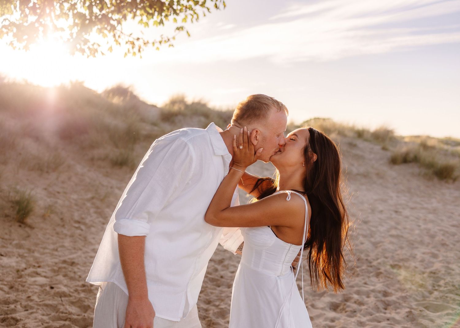 Couple sharing a romantic kiss on a sandy beach at golden sunset, wearing matching white outfits.