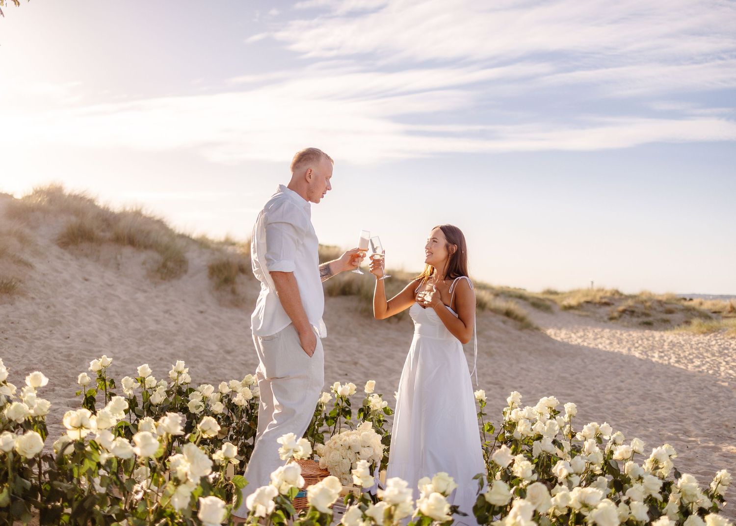Couple in white attire toasting champagne among white roses on a sandy beach at golden hour.