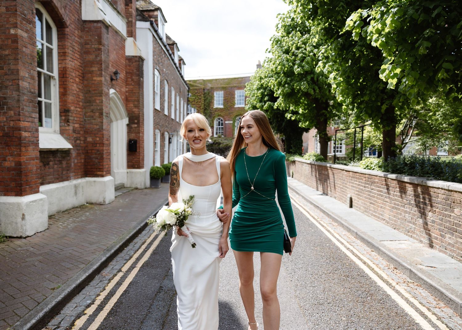 Bride in white gown and woman in green dress walking together on a sunny London street.