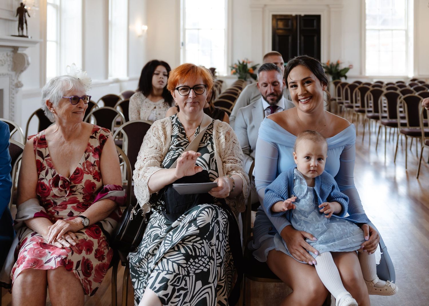 Guests seated at a wedding ceremony, including a woman in blue holding a baby, smiling warmly in an elegant venue.