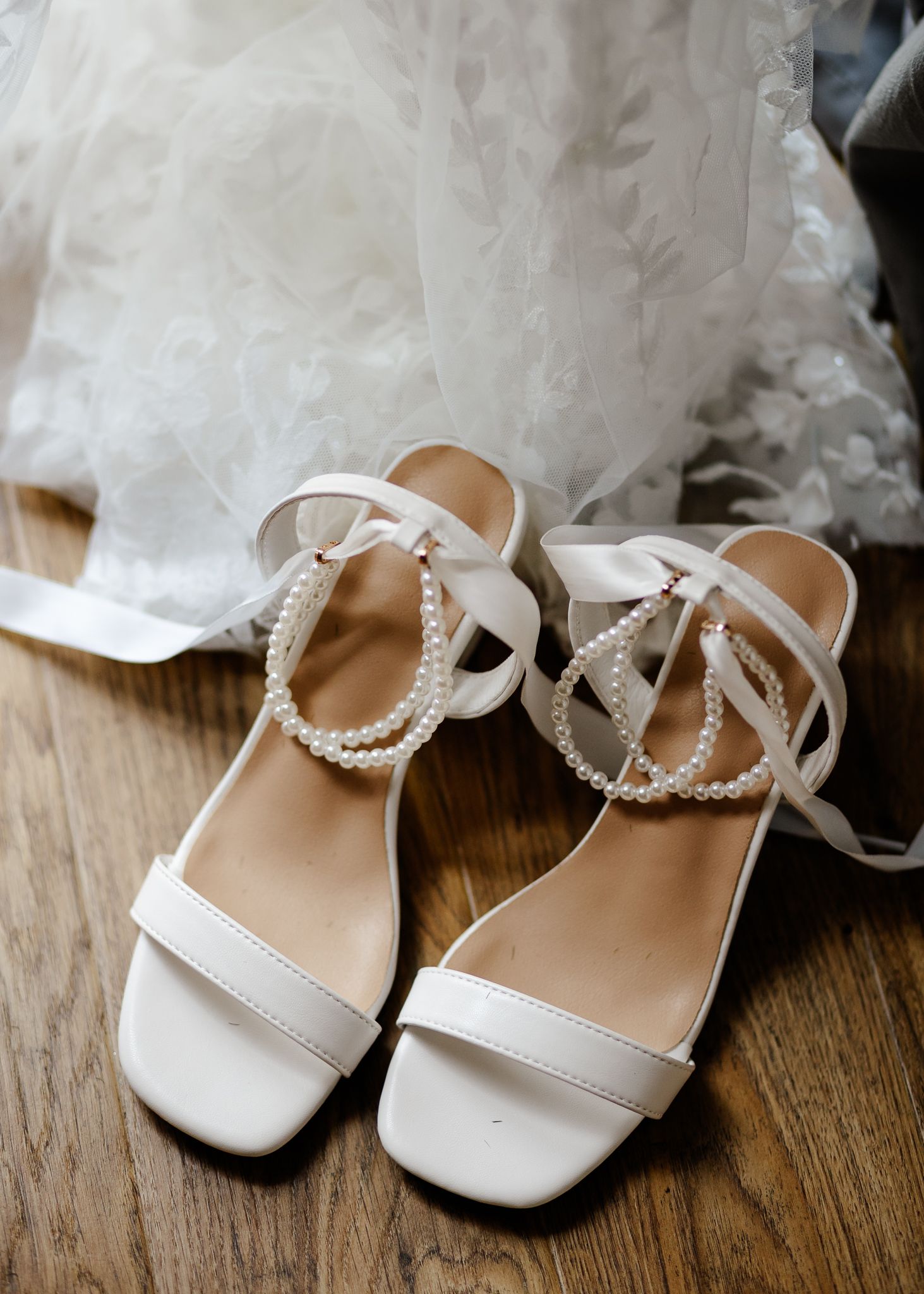 Elegant white satin wedding sandals with pearl straps displayed on a rustic wooden floor beside tulle fabric.