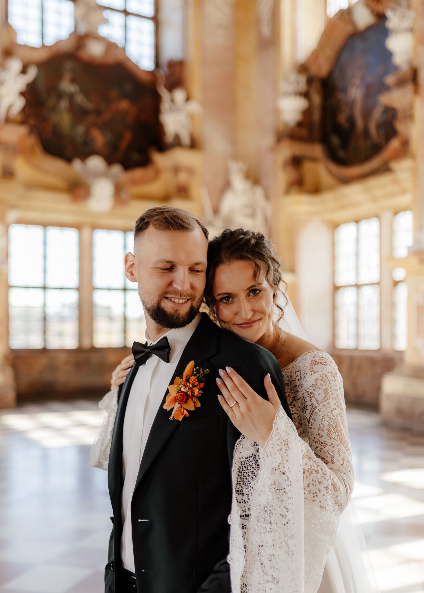 A romantic wedding couple embraces and smiles in a sunlit ornate hall with large windows.