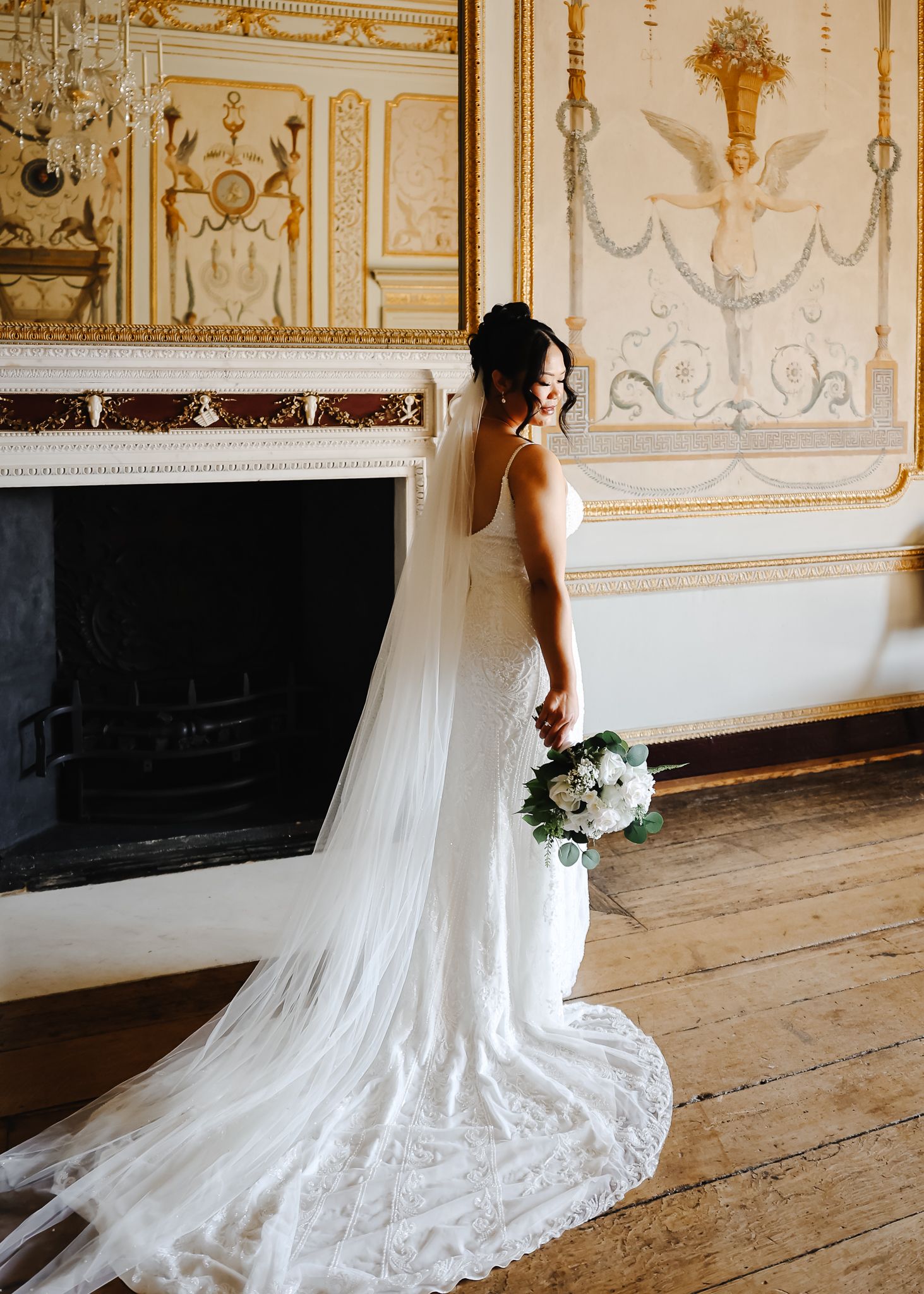 Elegant white wedding dress with flowing train displayed in an ornate room with gilded walls and wooden floors.