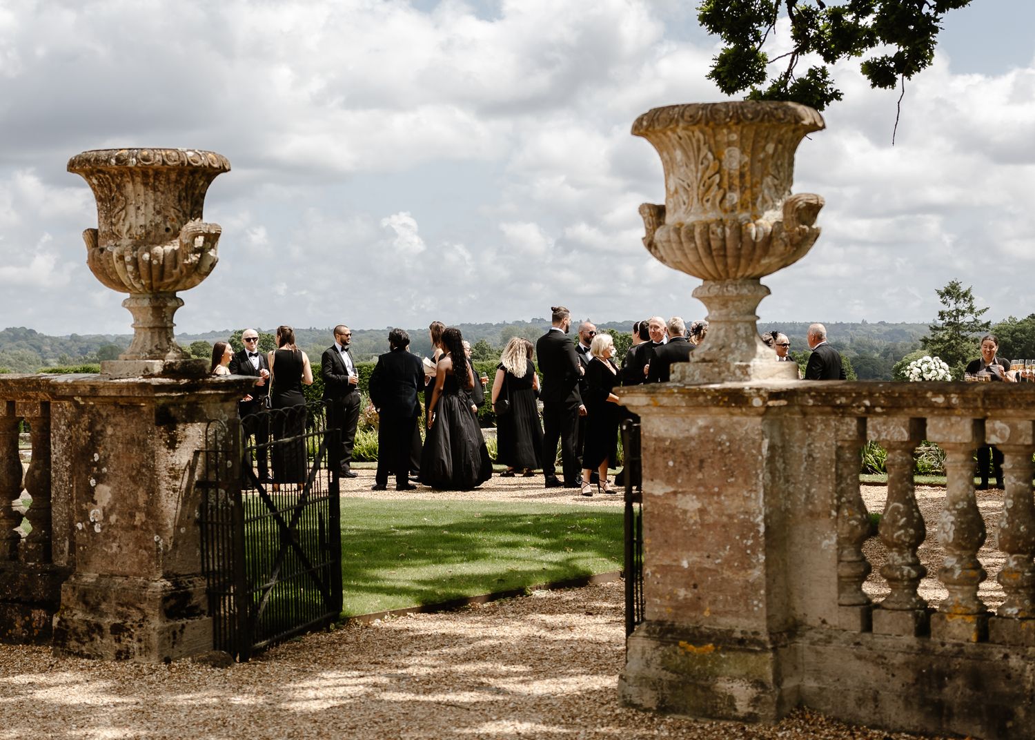 Ornate stone urns frame a garden entrance where wedding guests gather on a cloudy day.
