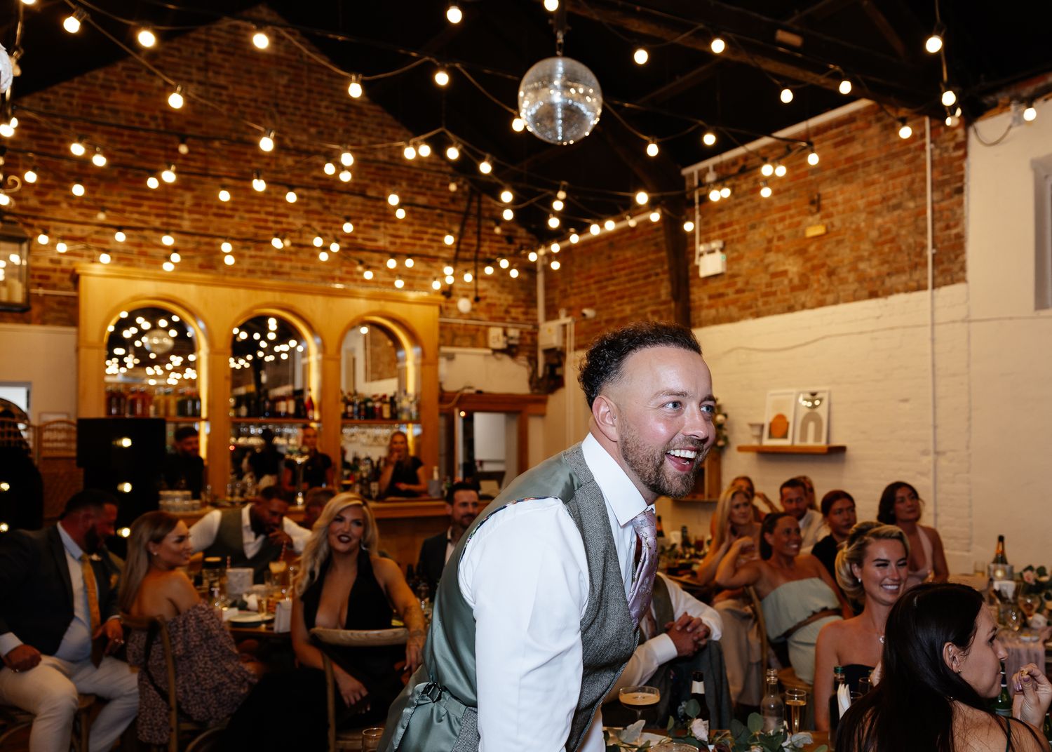 Wedding reception guests dancing and celebrating under string lights in a rustic brick venue space.