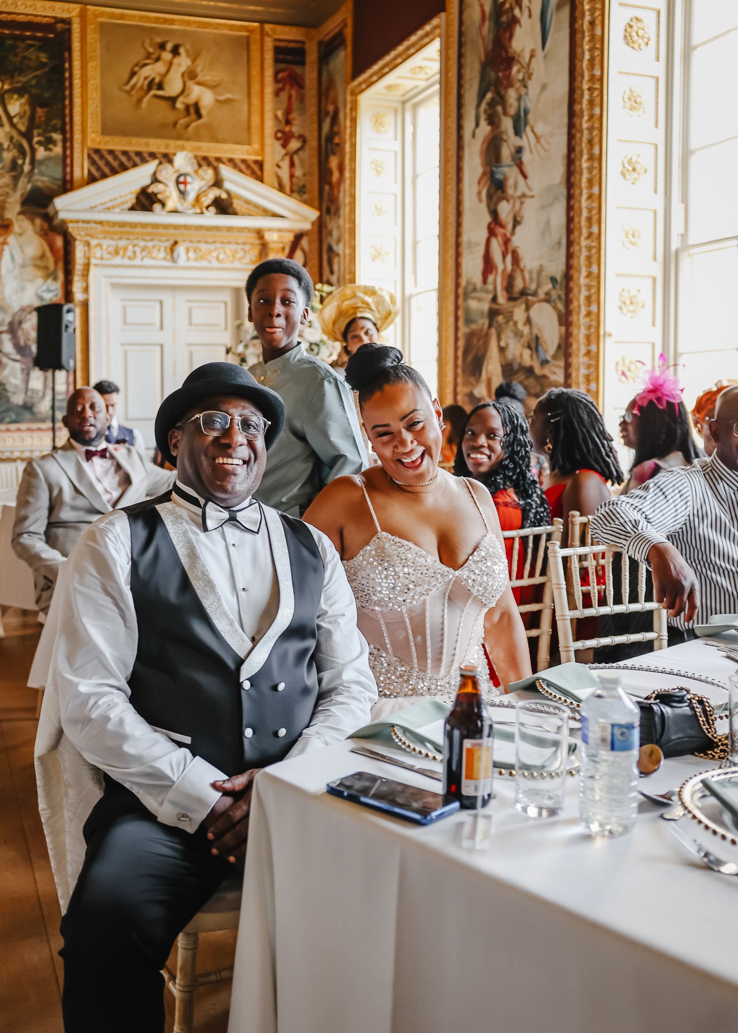 Guests celebrate joyfully at an elegant wedding reception in an ornate historic room with decorative wall paintings and windows.