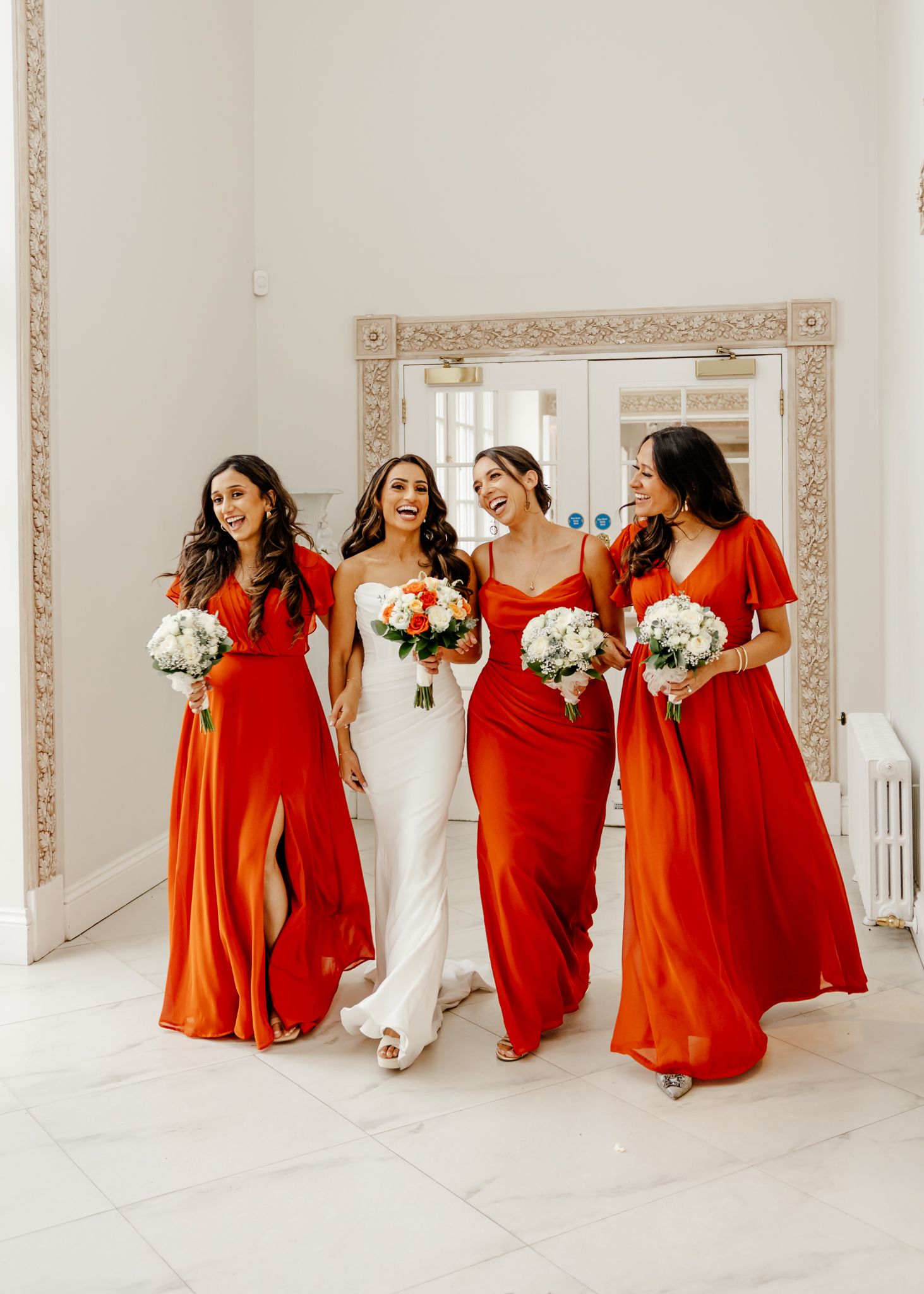 A bridal party in flowing red bridesmaid dresses and white wedding gown walk together in an elegant white room.