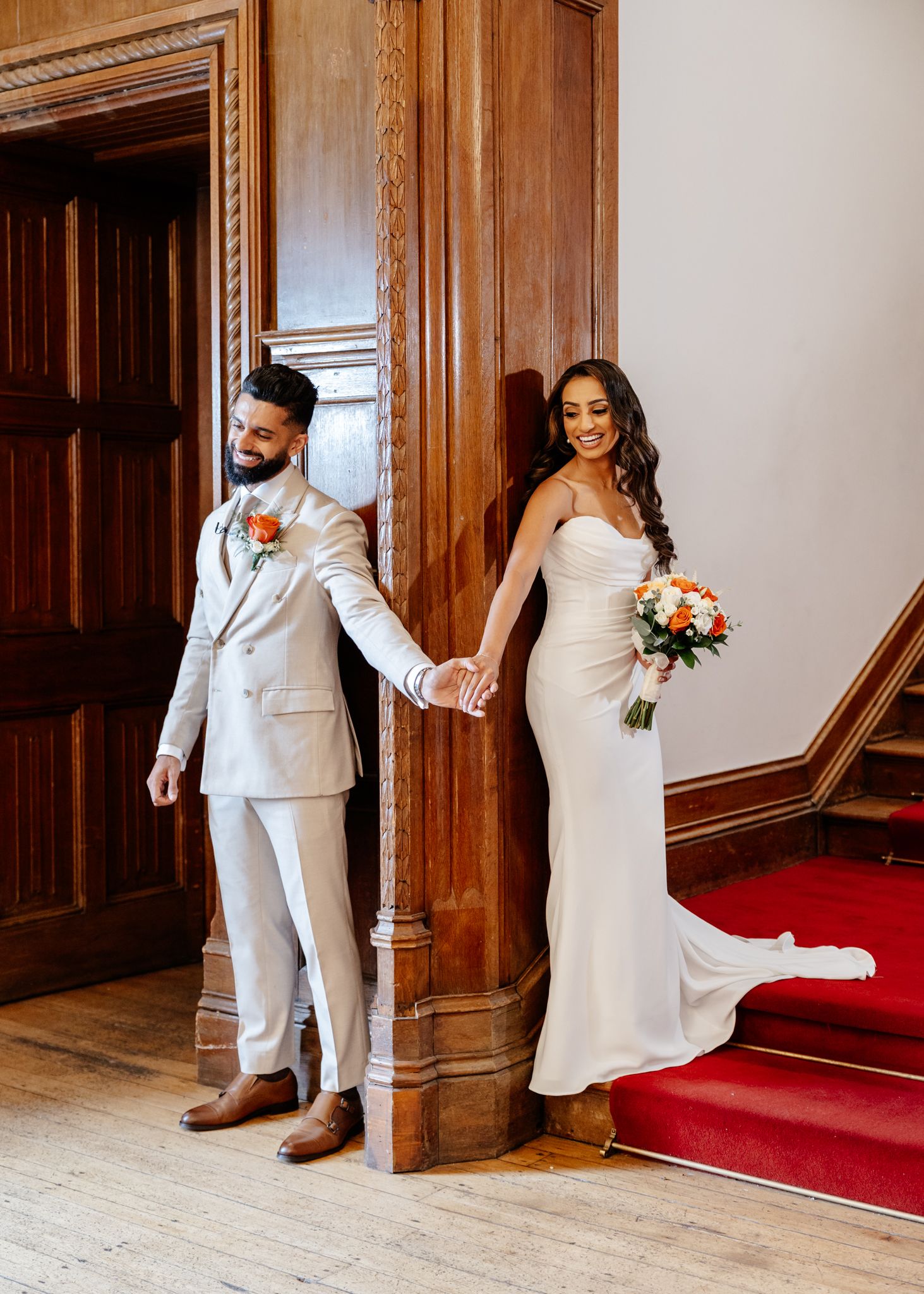 Wedding couple in white attire playfully pose behind wooden doors in an elegant venue with red carpeting.