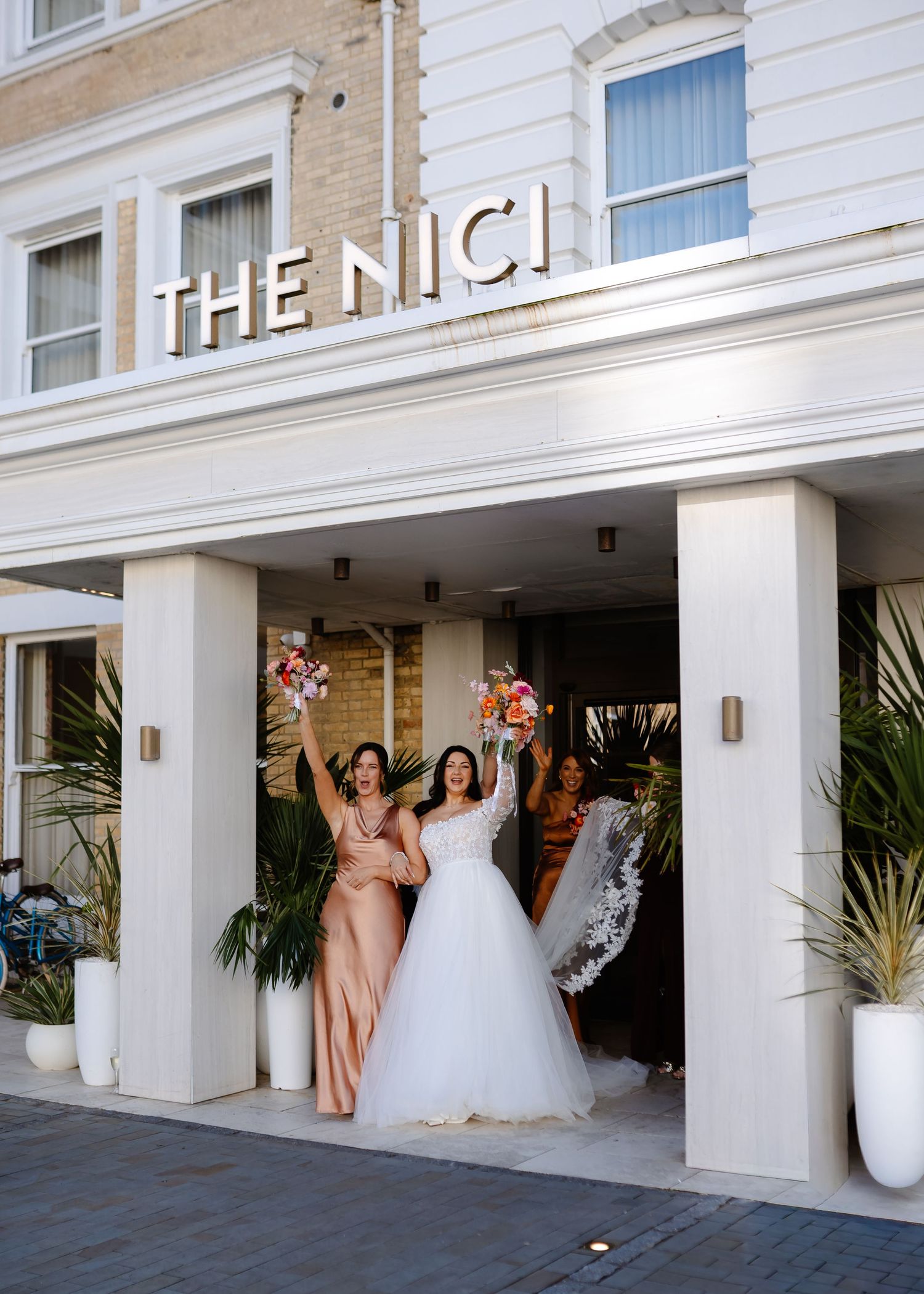 Bride and bridesmaid celebrate outside The Nici hotel, raising colorful bouquets in wedding attire.
